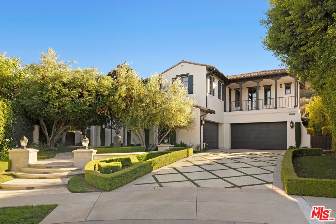 Two-story stucco home with a tiled roof, balcony, geometric driveway, and formal front garden.