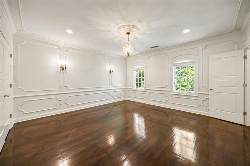 Empty formal room with ornate white wall paneling, dark hardwood floors, and a crystal chandelier.