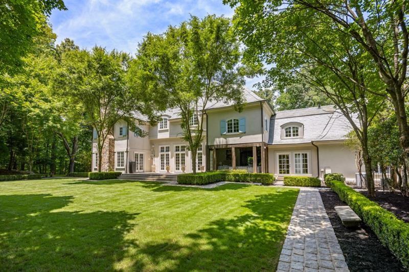 Large light-colored home with blue shutters, mature trees, and a stone walkway across the lawn.