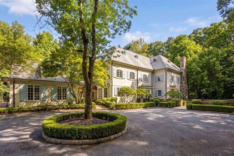 Large two-story house framed by mature trees, with a circular driveway planter and clipped hedges.