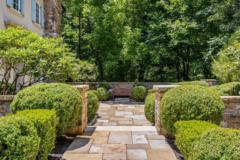 Stone garden walkway bordered by trimmed hedges leads to a wooden bench beside the house.