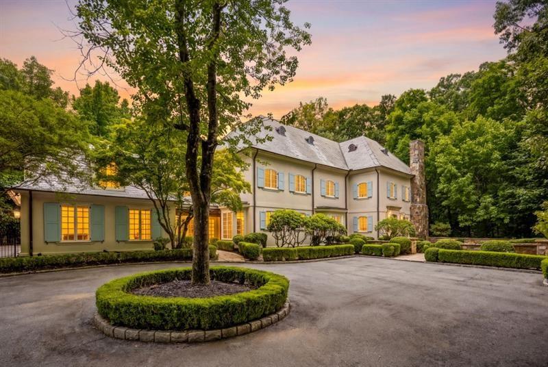 Large two-story house with an attached wing, manicured hedges, and a circular driveway tree bed at dusk.