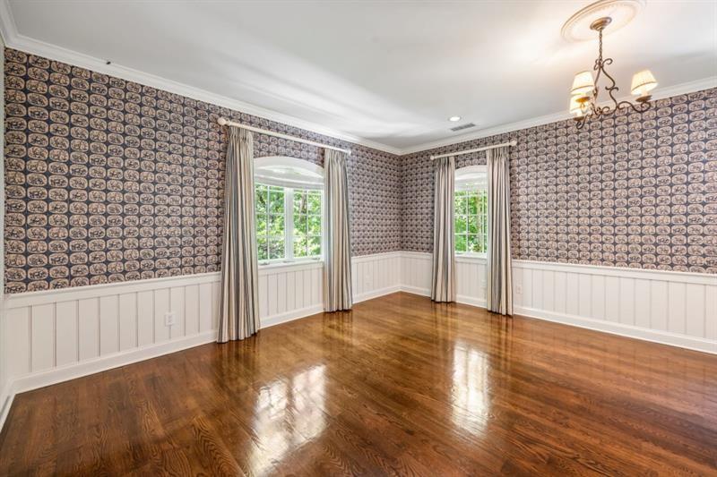 Empty room with patterned wallpaper, white wainscoting, hardwood floors, and two curtained windows.