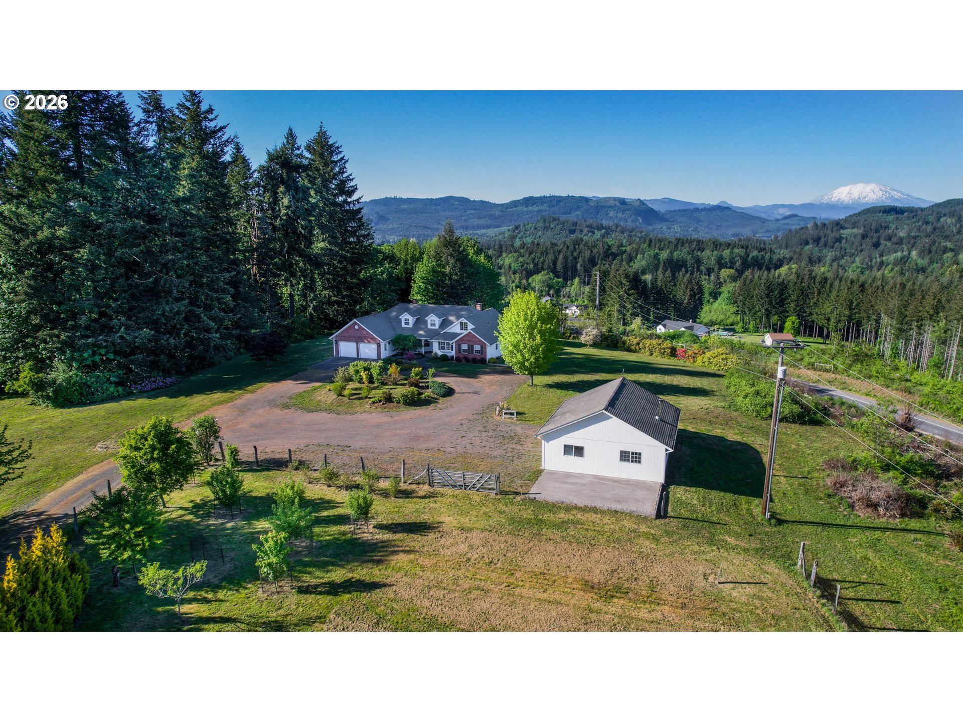 A rural home and detached outbuilding amid broad lawns, evergreen forest, and a distant snow-capped mountain.
