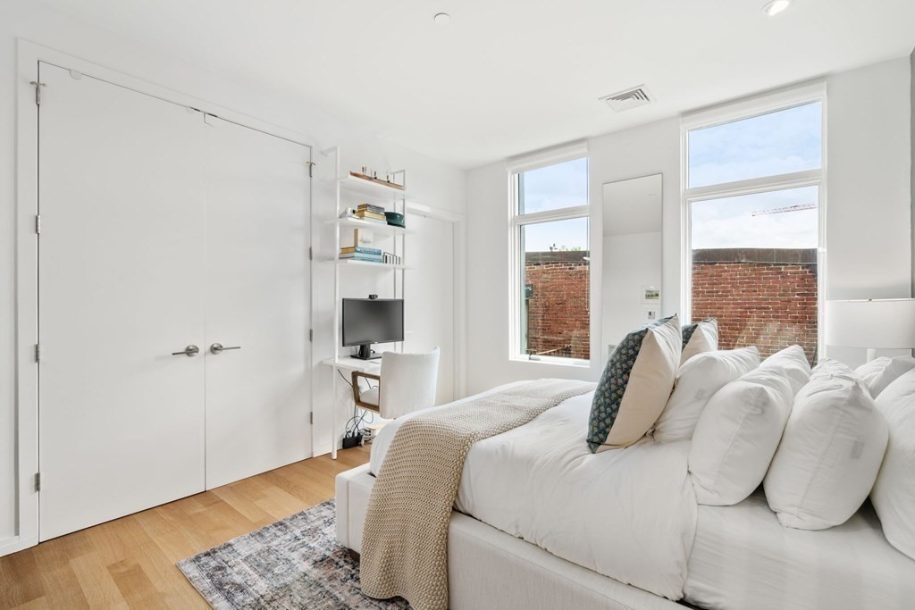 Bright bedroom with a white bed, two tall windows, and a compact desk and shelving area.