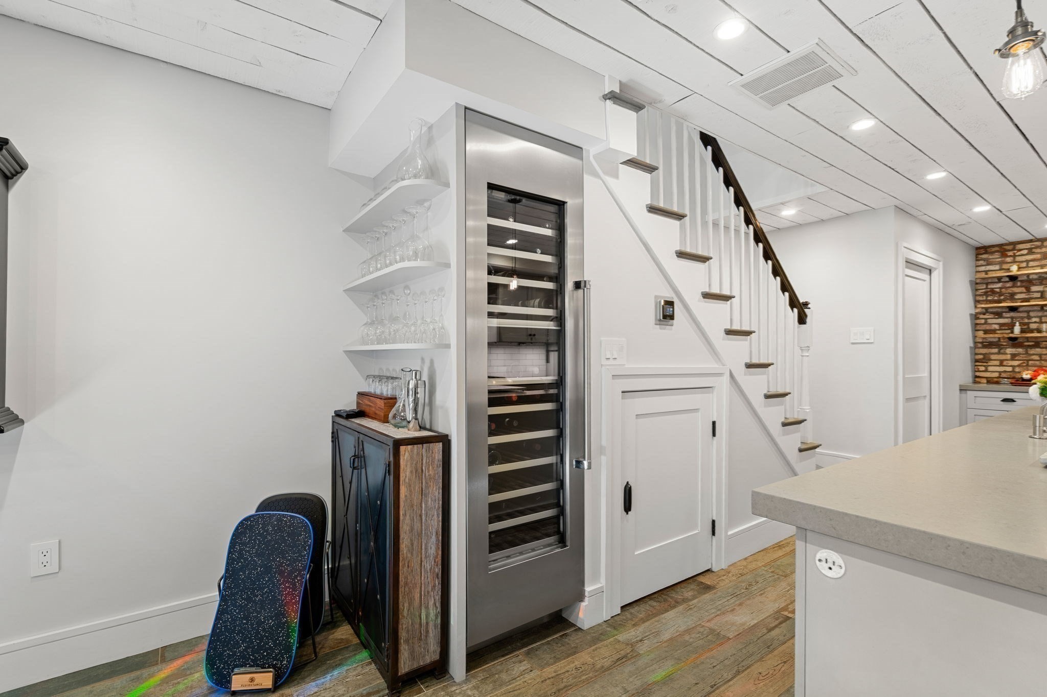Kitchen with a stainless-steel wine refrigerator, staircase, white island, and brick backsplash.