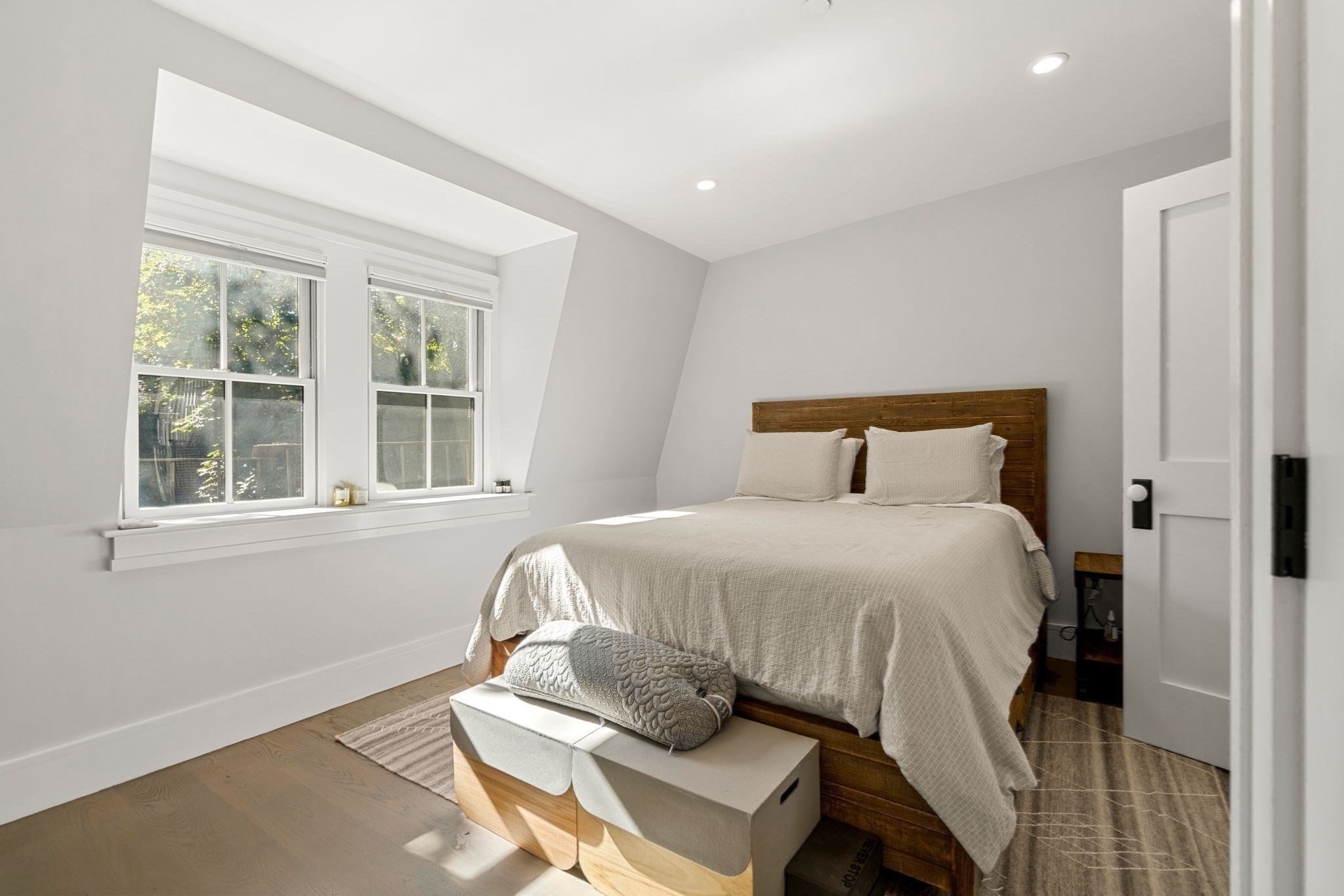 Bedroom with a wooden bed, pale bedding, and two sunlit windows.