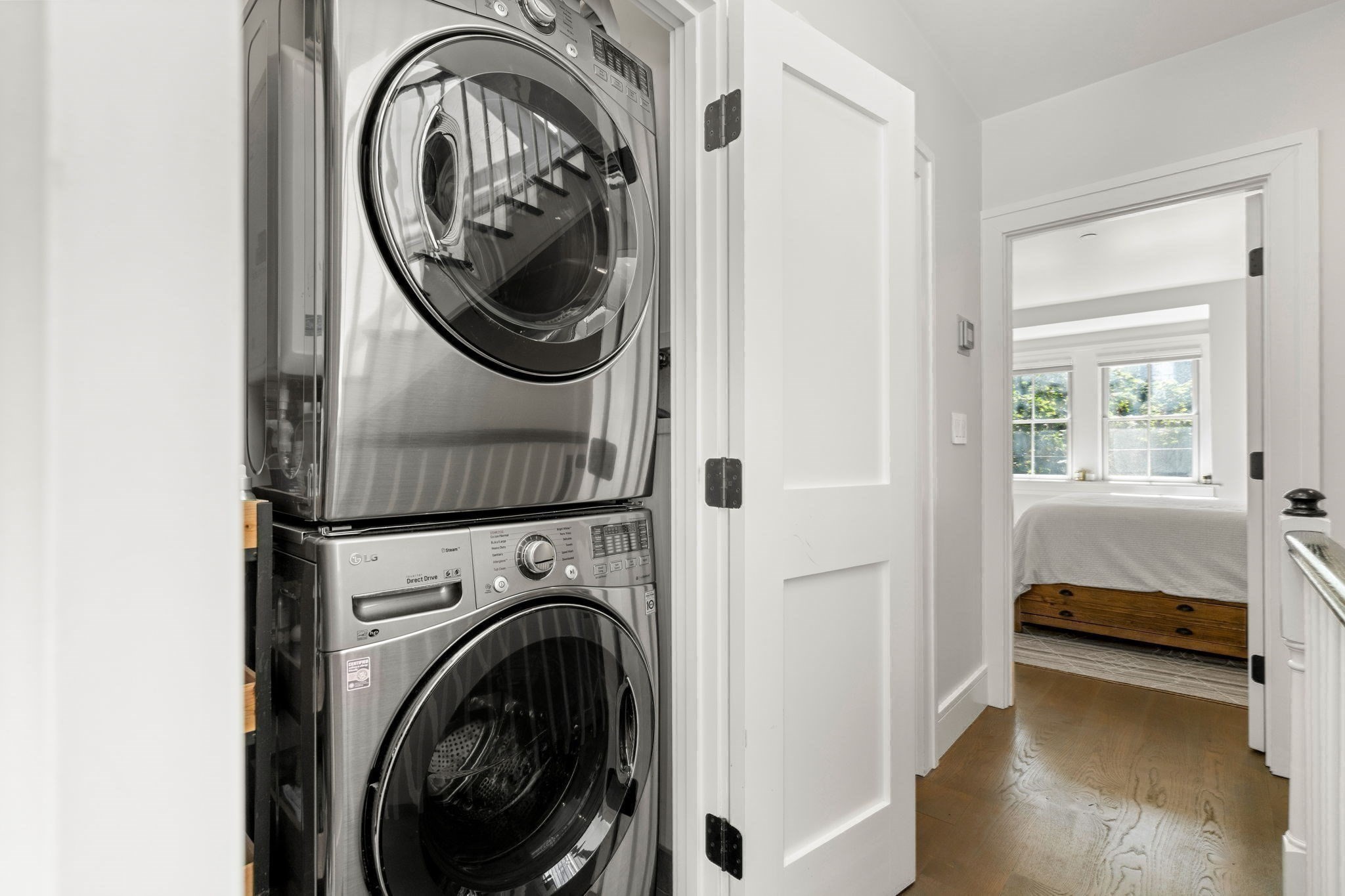 Stacked stainless-steel washer and dryer beside an open doorway to a bright bedroom.