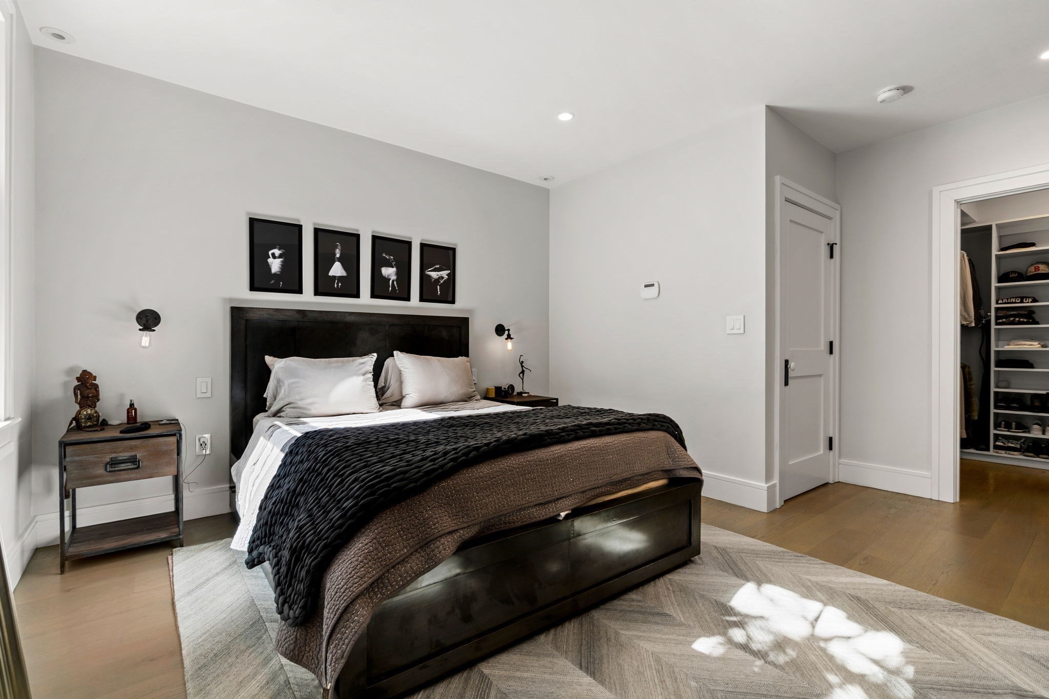 Modern bedroom with a dark bed, layered bedding, framed prints, and an open walk-in closet.