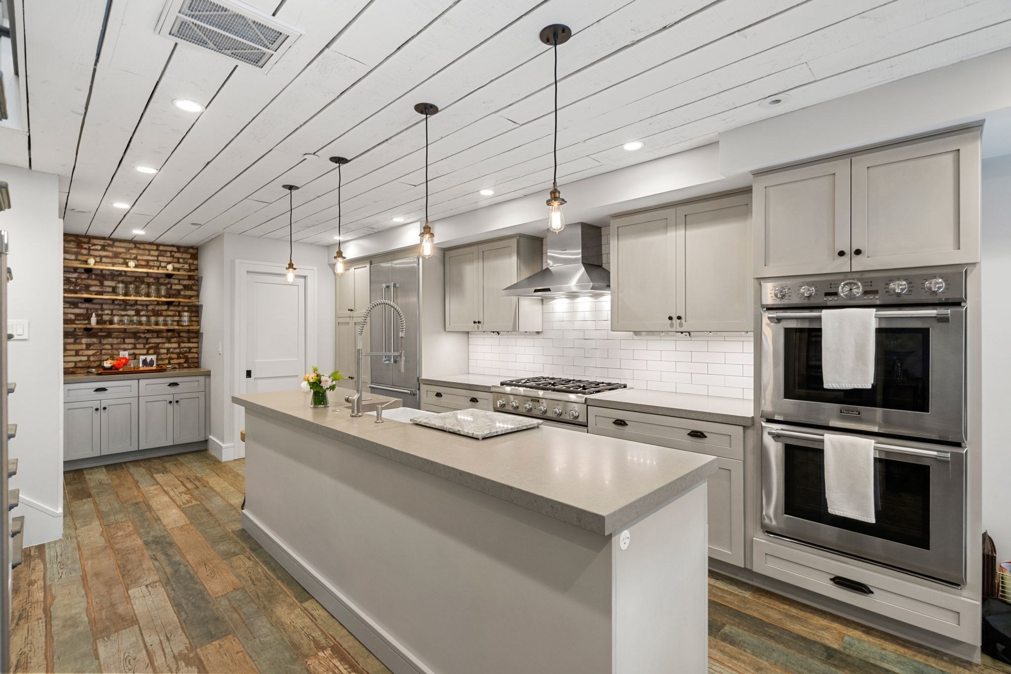 Kitchen with a large island, gray cabinets, stainless steel double ovens, and exposed brick shelving.