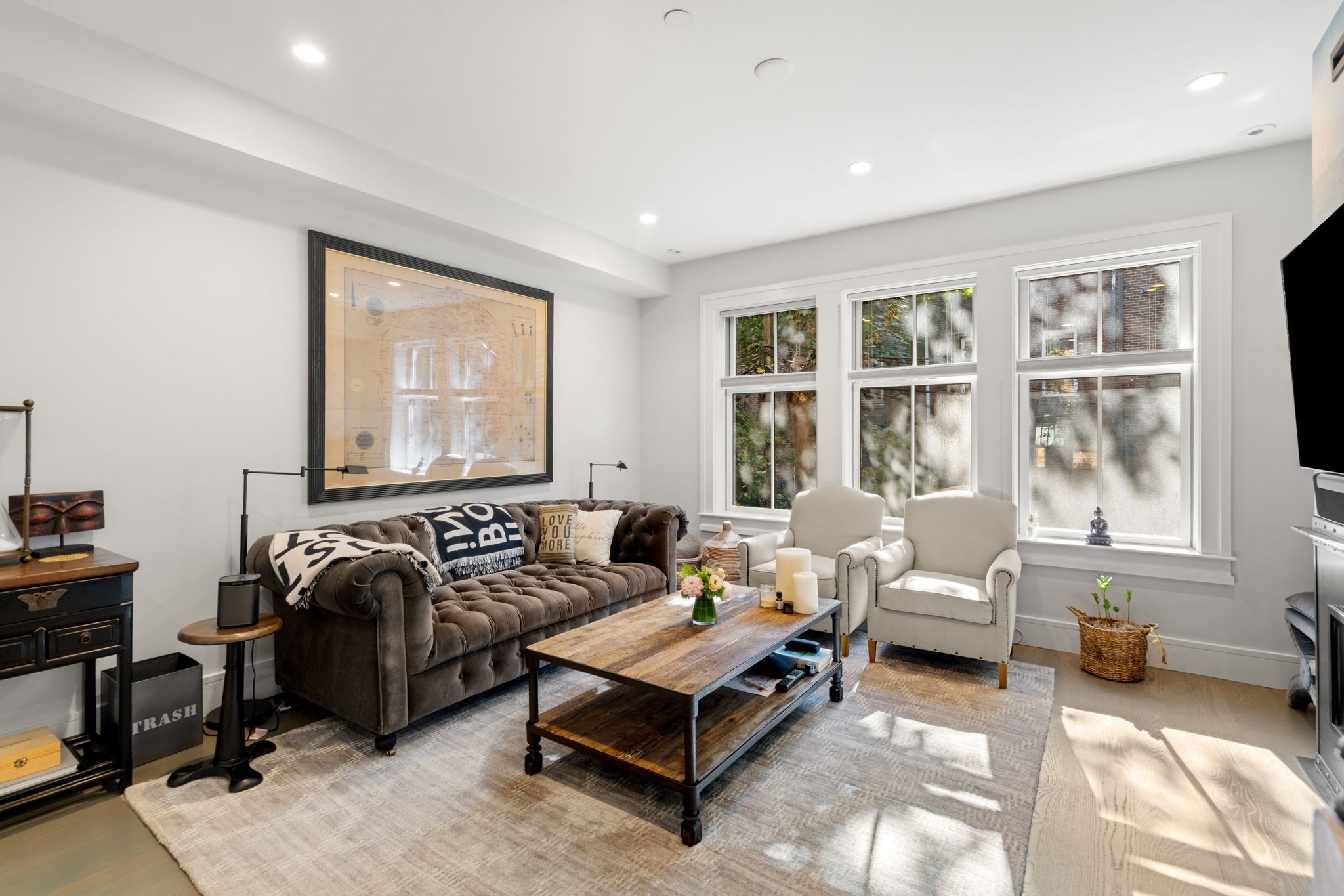 Living room with a tufted brown sofa, two white armchairs, and three large sunlit windows.