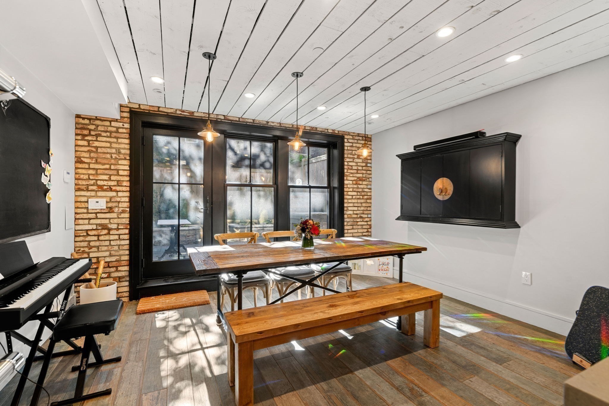 Dining room with exposed brick, black-framed glass doors, and a long rustic wood table.