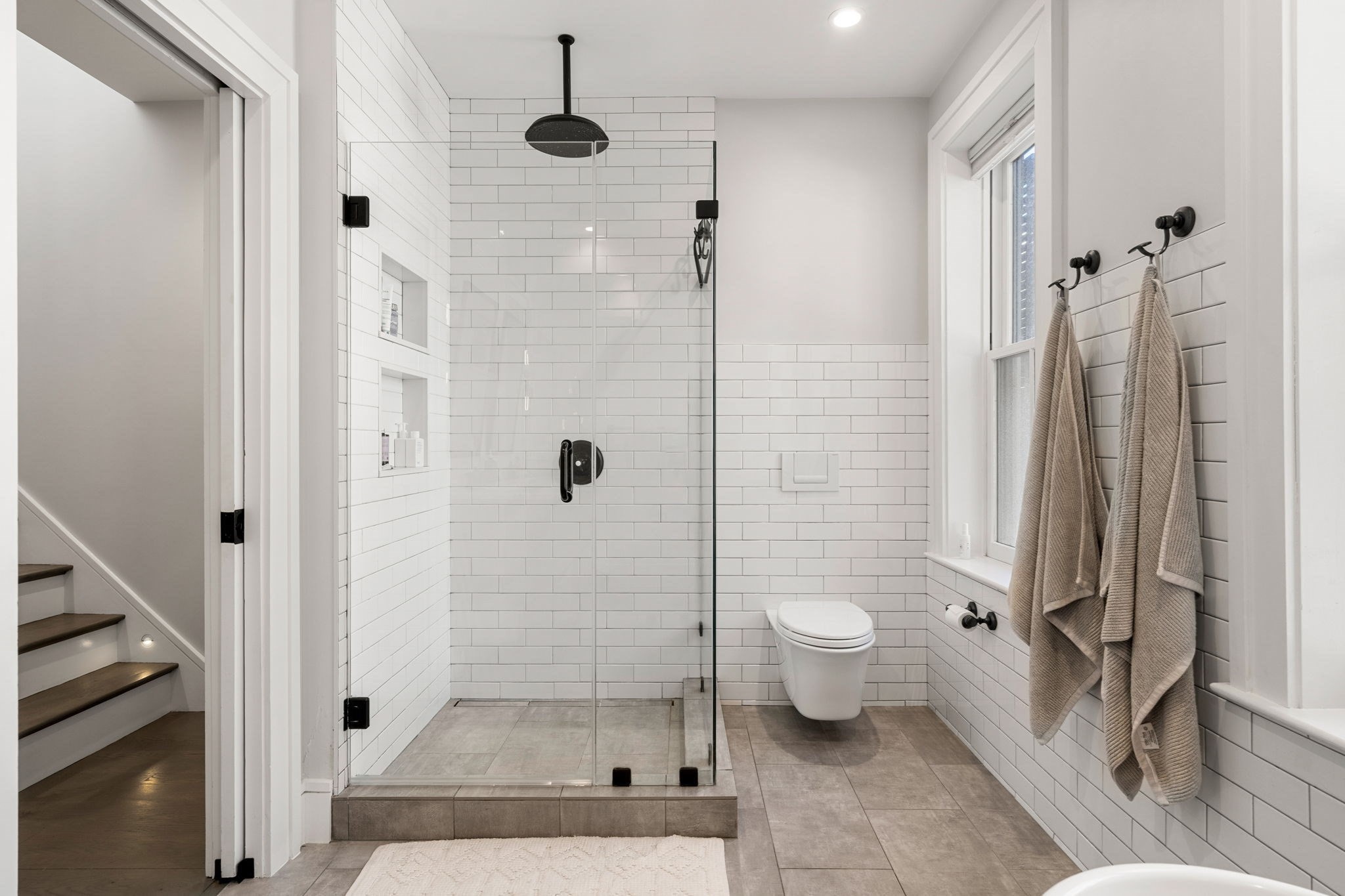 Bathroom with white subway tile, glass walk-in shower, black rain showerhead, and beige towels beside a window.