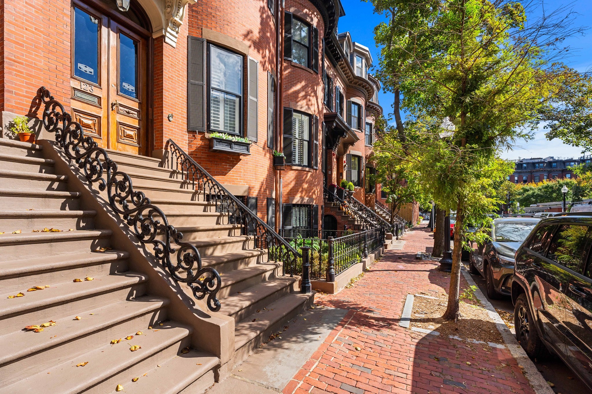 Red-brick Boston townhouse with broad stone steps and ornate iron railings along a tree-lined brick sidewalk.