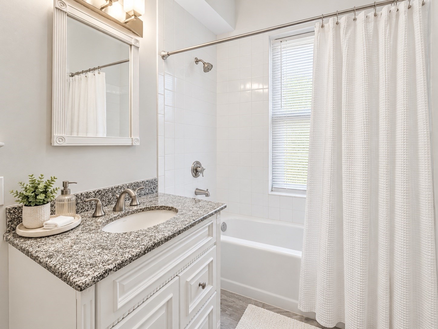 Bathroom with a granite vanity and white tiled tub-shower beside a curtained window.