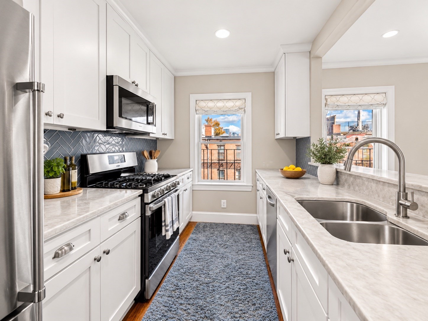 Renovated galley kitchen with white cabinets, marble counters, stainless steel appliances, and windows overlooking brick buildings.