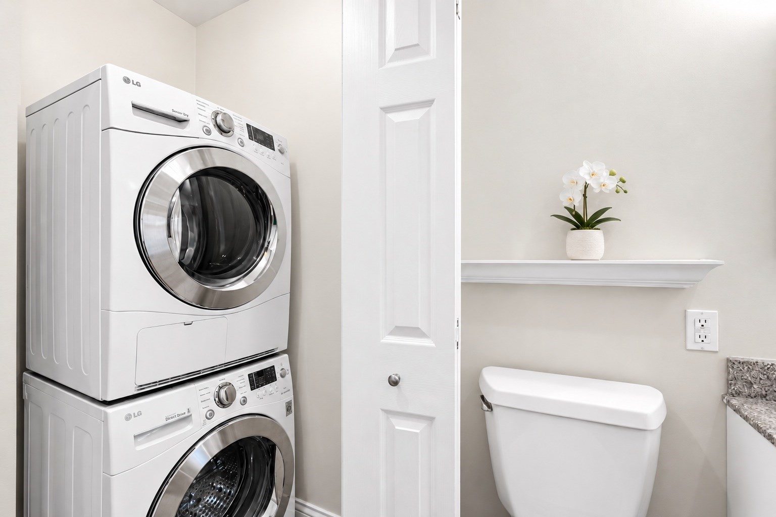 Compact bathroom laundry area with stacked front-loading machines, a toilet, and an orchid on a wall shelf.