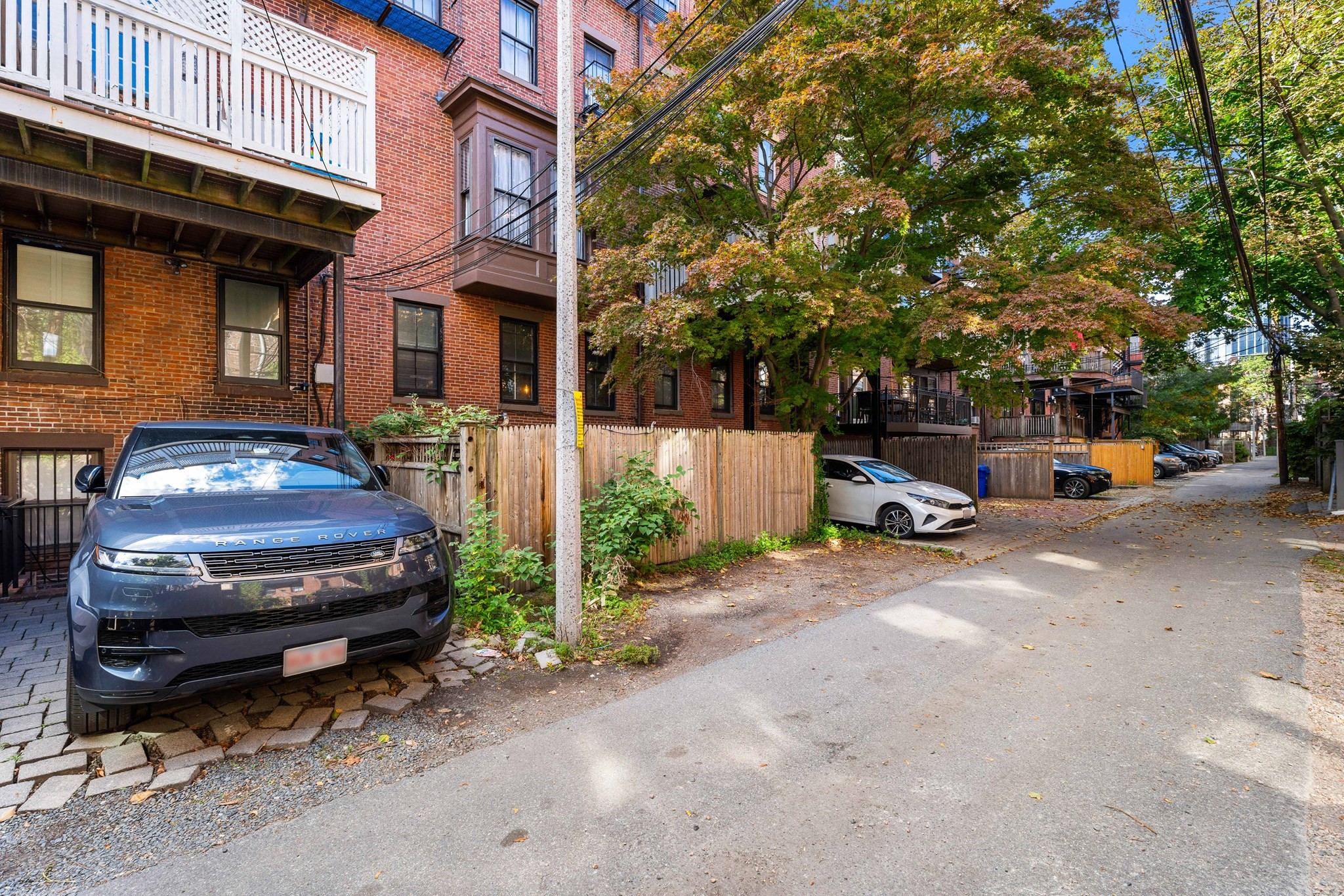 Red-brick residential buildings and fenced parking along a tree-lined lane, with a dark SUV in the foreground.