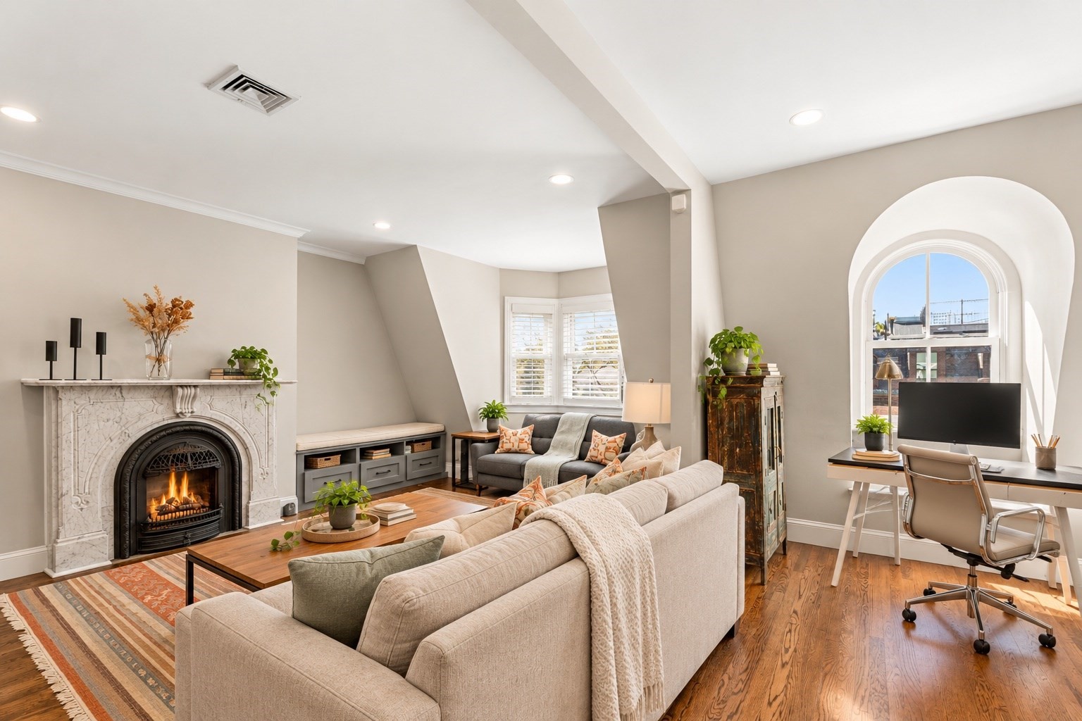 Staged living room with a marble fireplace, neutral sofas, and a desk beneath an arched window.