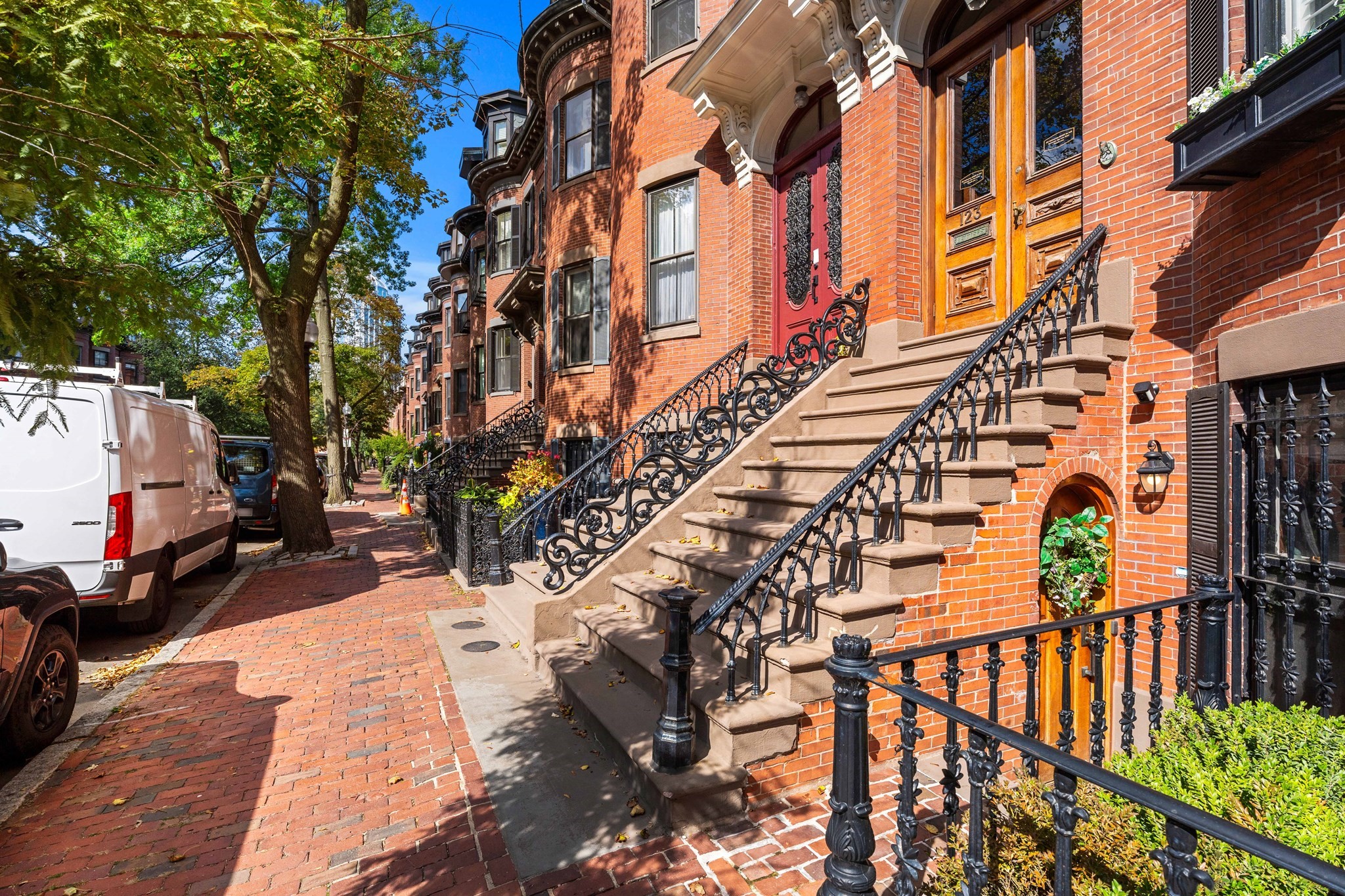 Red-brick townhouse with a raised stone stoop, ornate iron railings, and a tree-lined brick sidewalk.