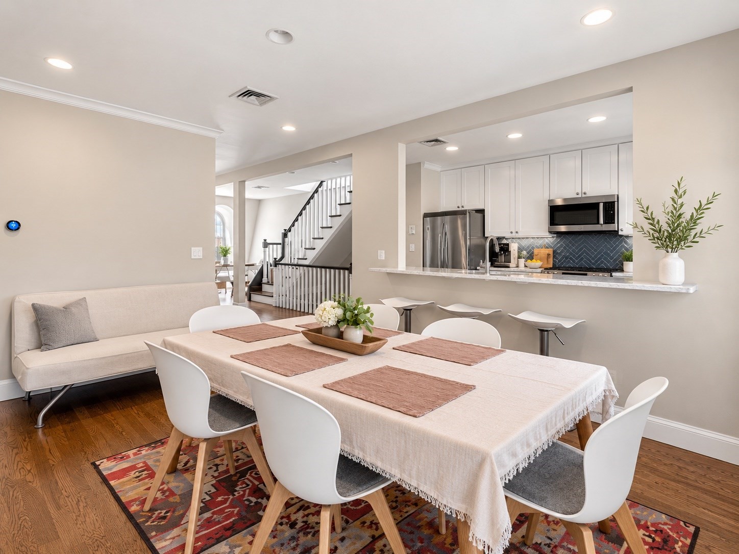 Dining area with a long table and white chairs beside an open kitchen with blue tile backsplash.