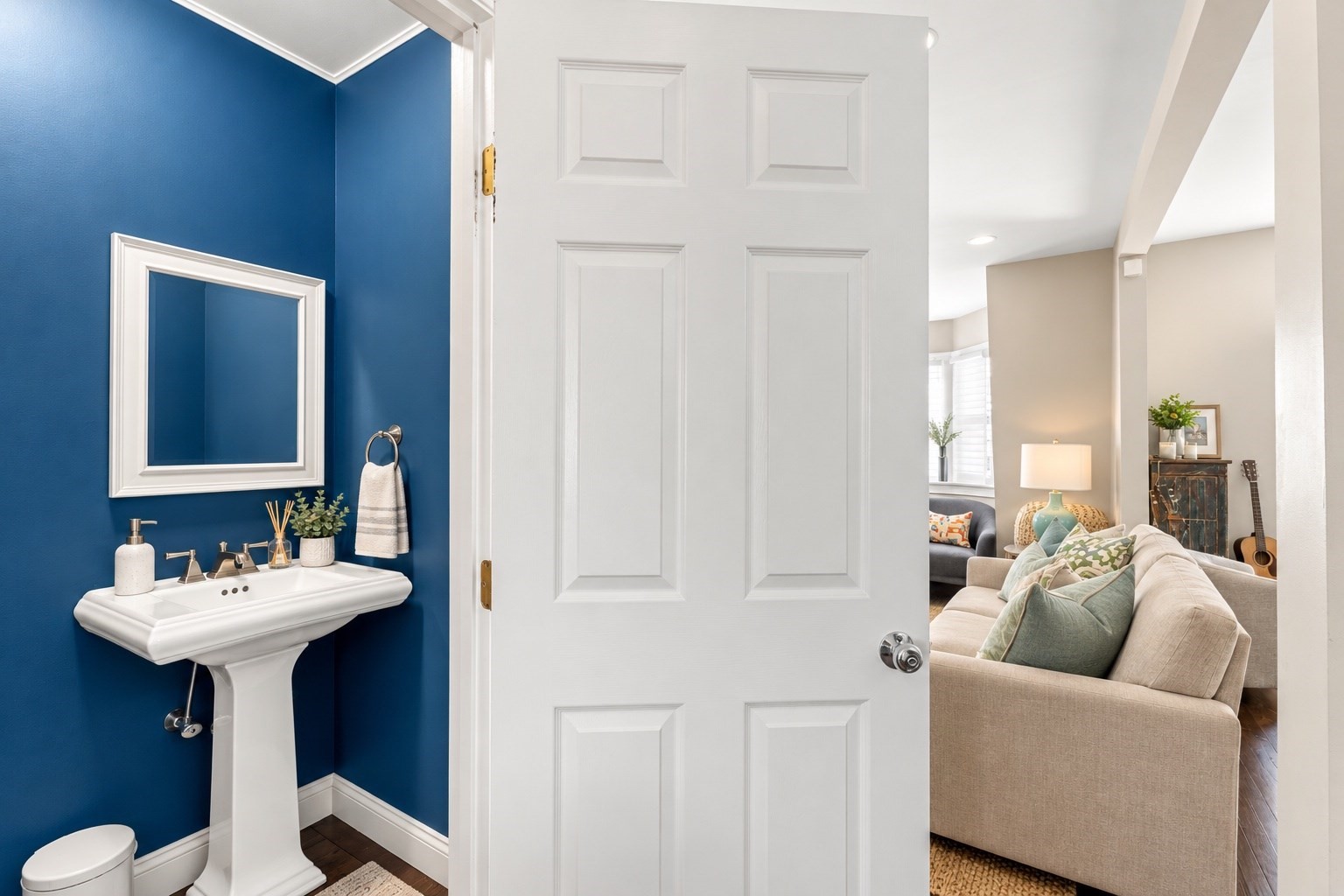 Blue powder room with pedestal sink opens onto a living room with a beige sofa.
