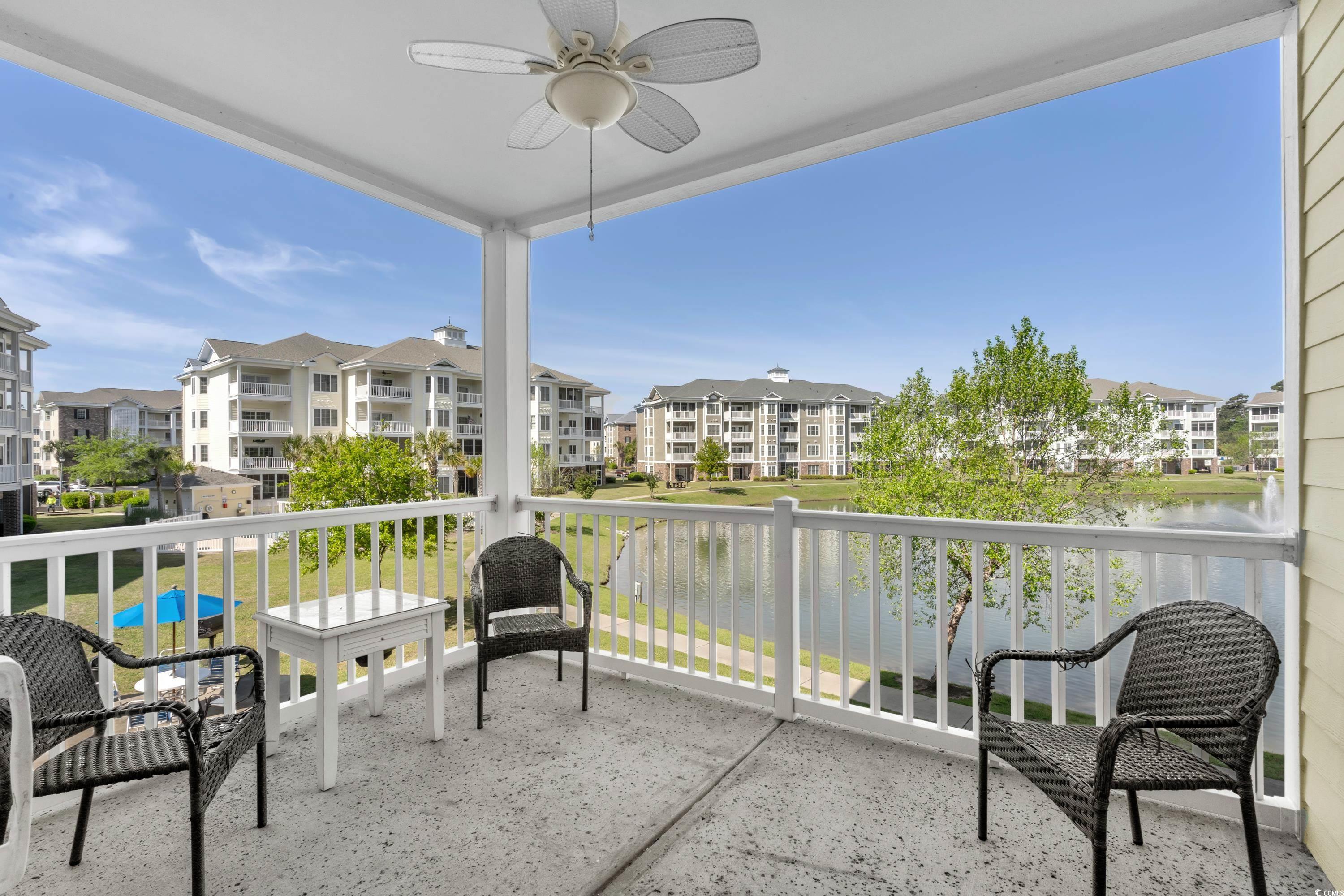 Covered balcony with wicker chairs overlooking a pond and neighboring apartment buildings.