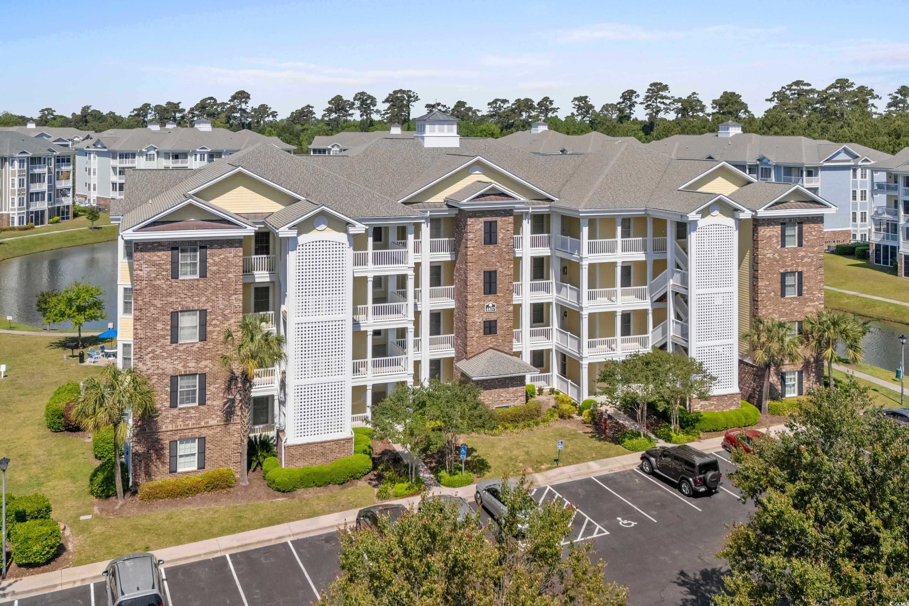 Aerial view of the 4837 condominium building, with white balconies, landscaped grounds, and a pond nearby.