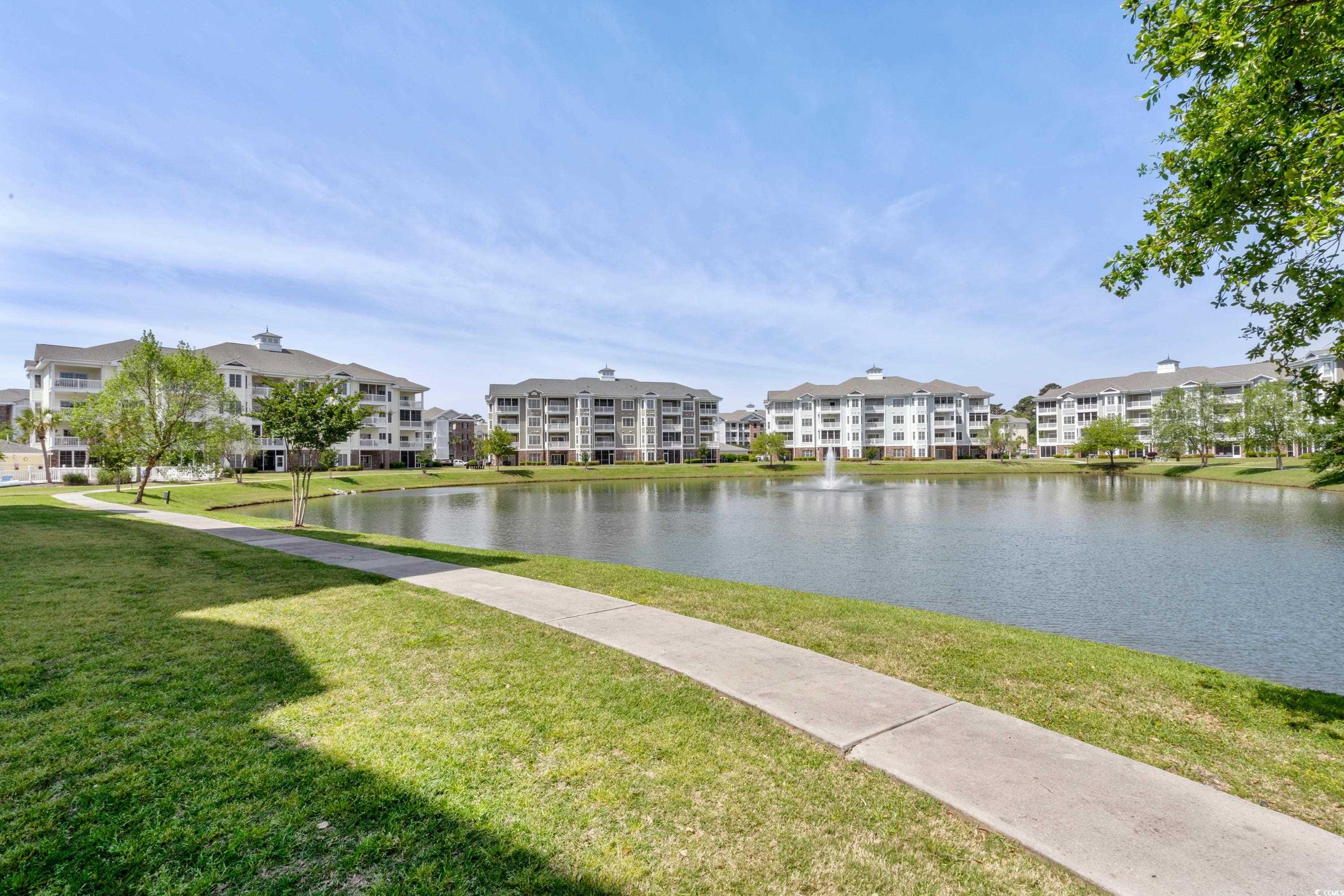 Condominium buildings across a pond with a fountain and paved walking path.