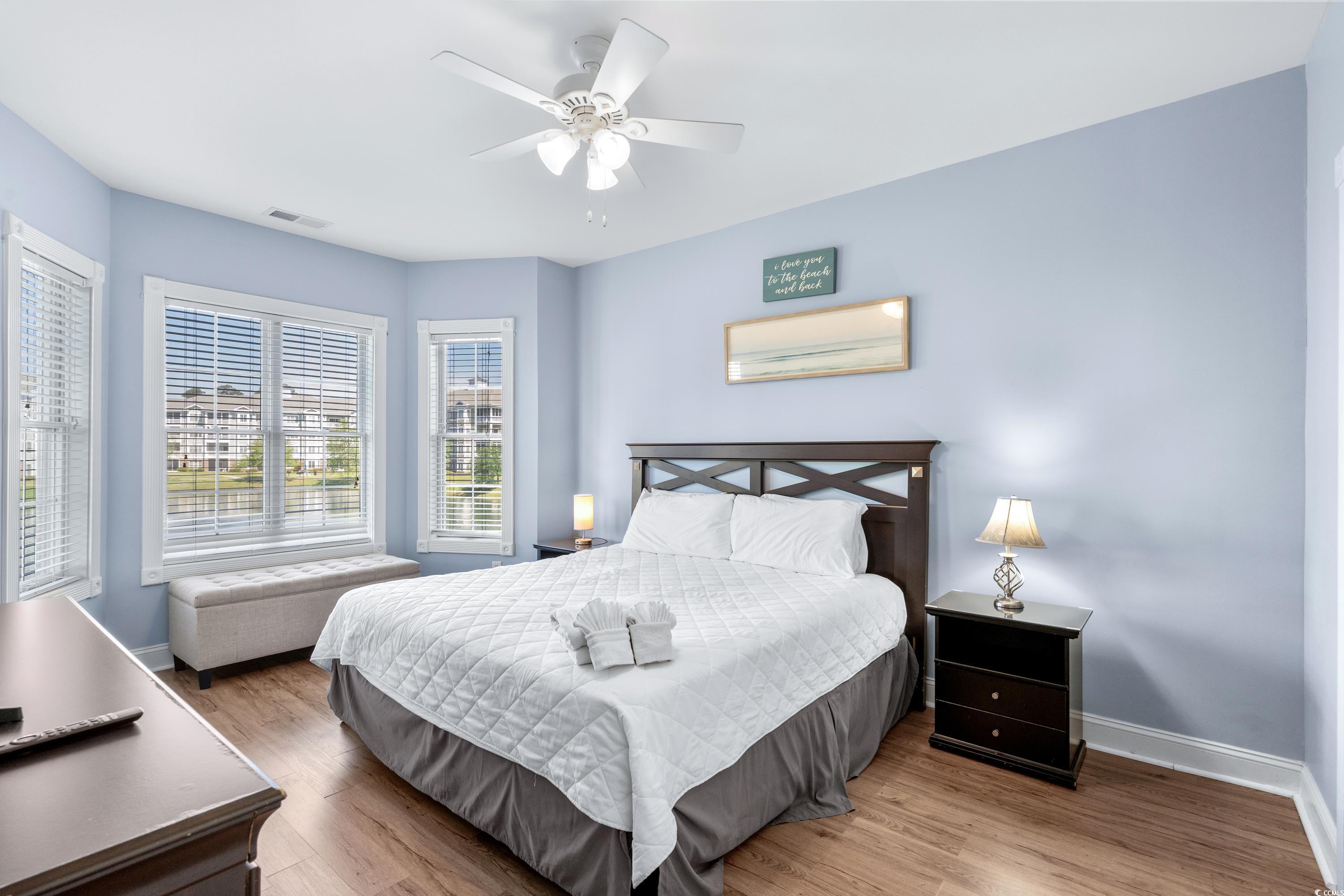 Bedroom with a white-quilted king bed, pale blue walls, and multiple windows overlooking water and nearby buildings.