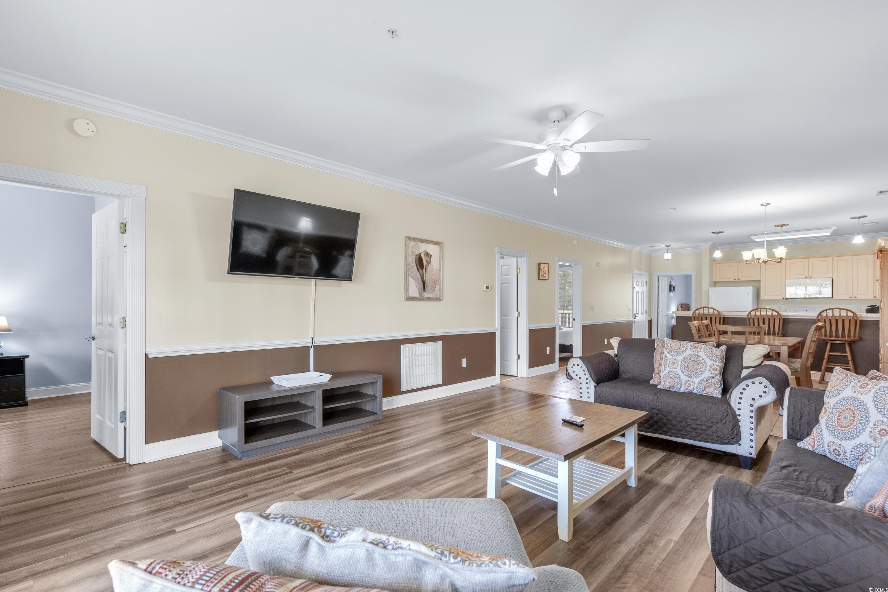 Open-plan living room with gray sofas, wall-mounted TV, wood-look floors, and dining area beside the kitchen.