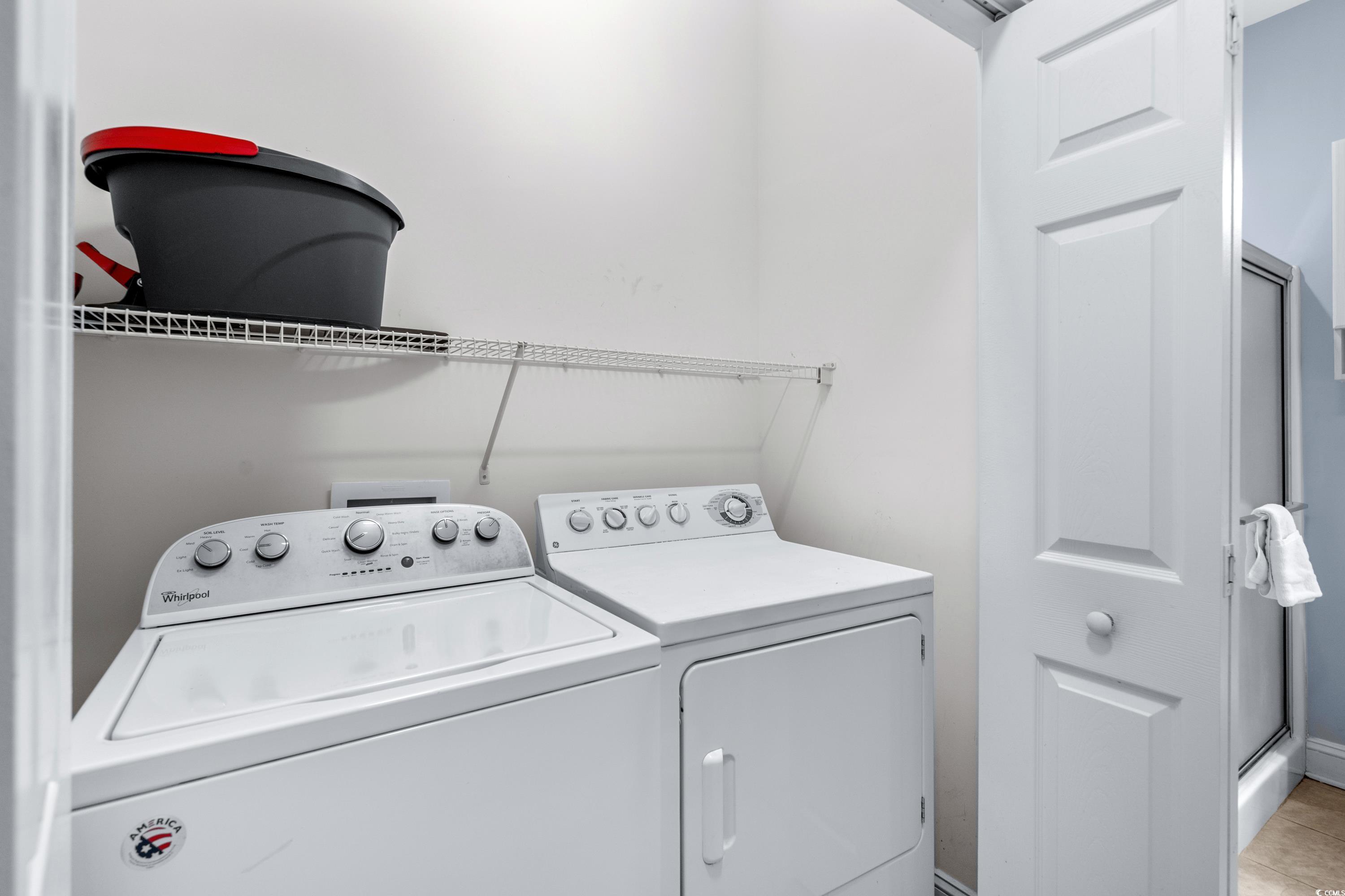 Laundry room with a white washer and dryer beneath a wire shelf holding a black-and-red storage bin.