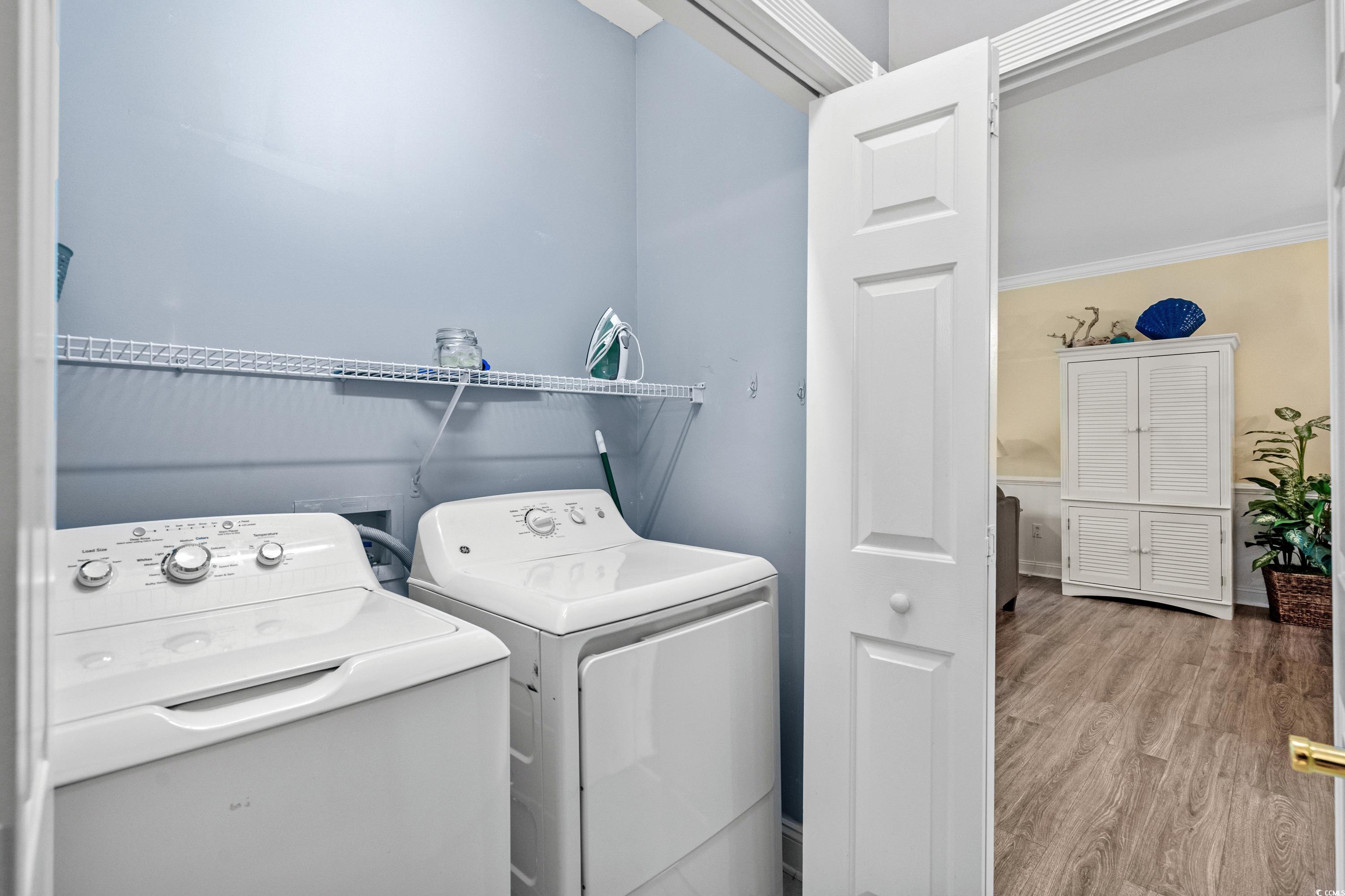Laundry room with washer and dryer beneath a wire shelf, opening to a furnished living area.