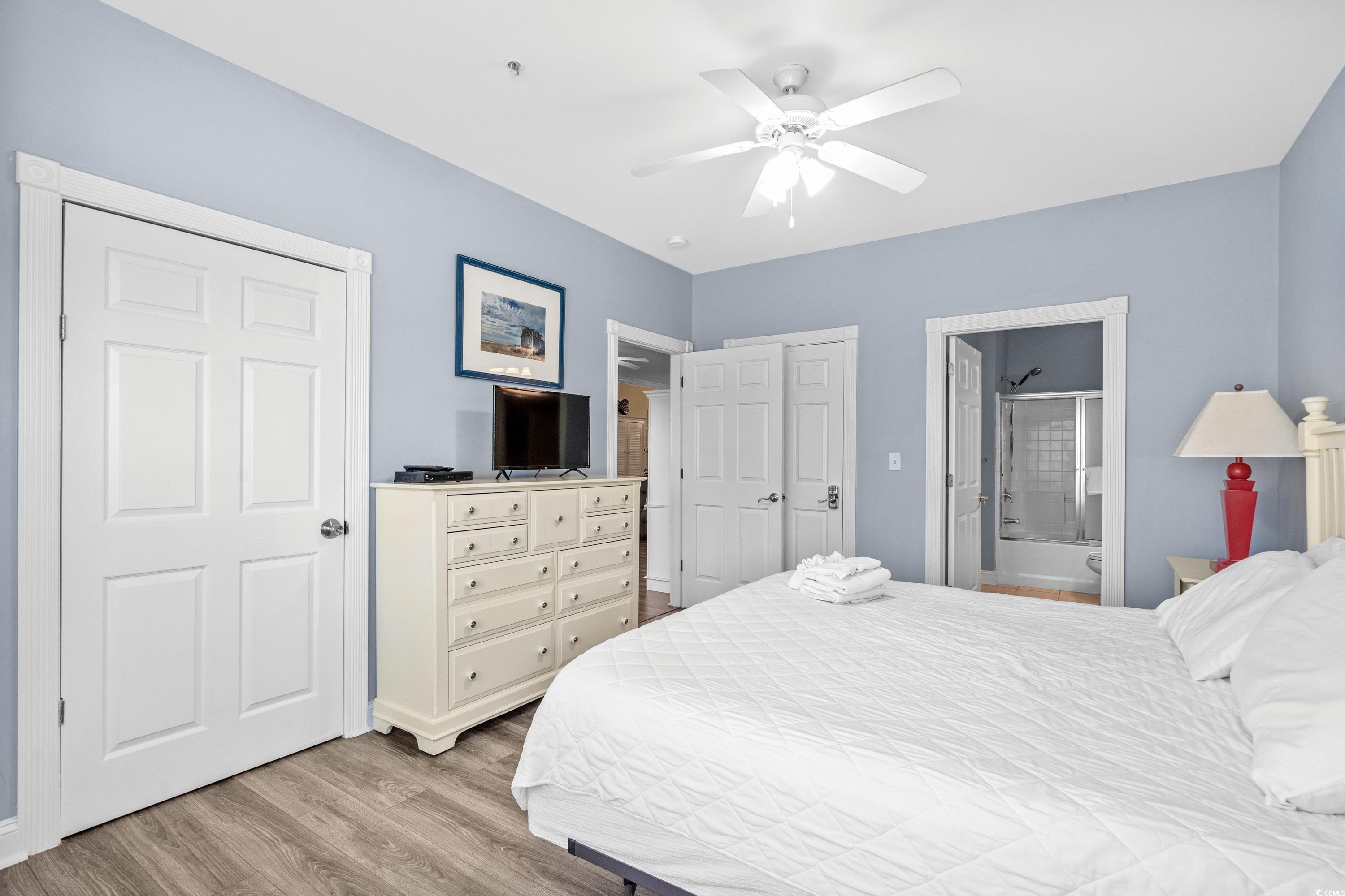 Bedroom with a white bed, pale blue walls, dresser, ceiling fan, and open bathroom doorway.