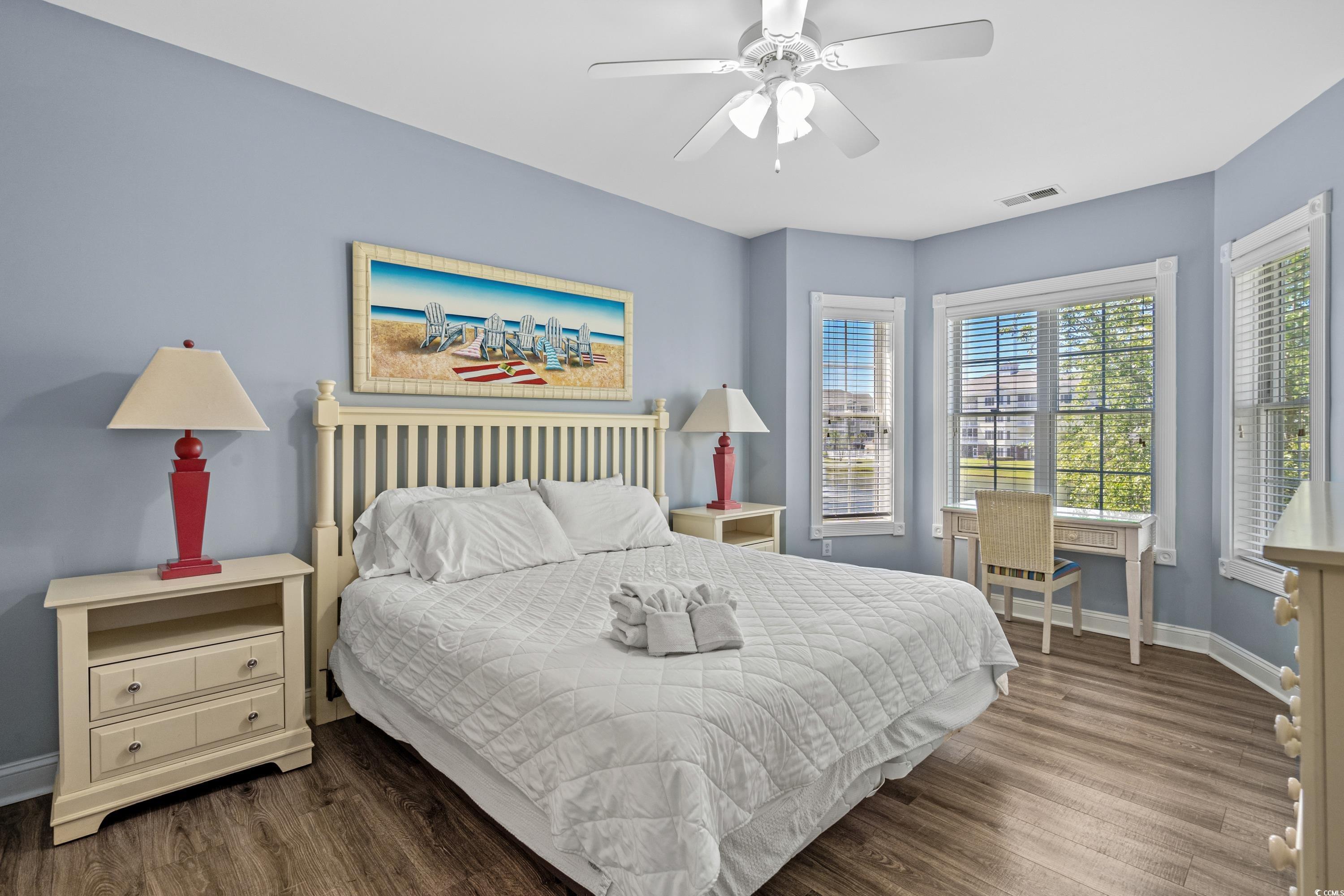 Bedroom with a white king bed, beach-chair wall art, pale blue walls, and three bright windows.