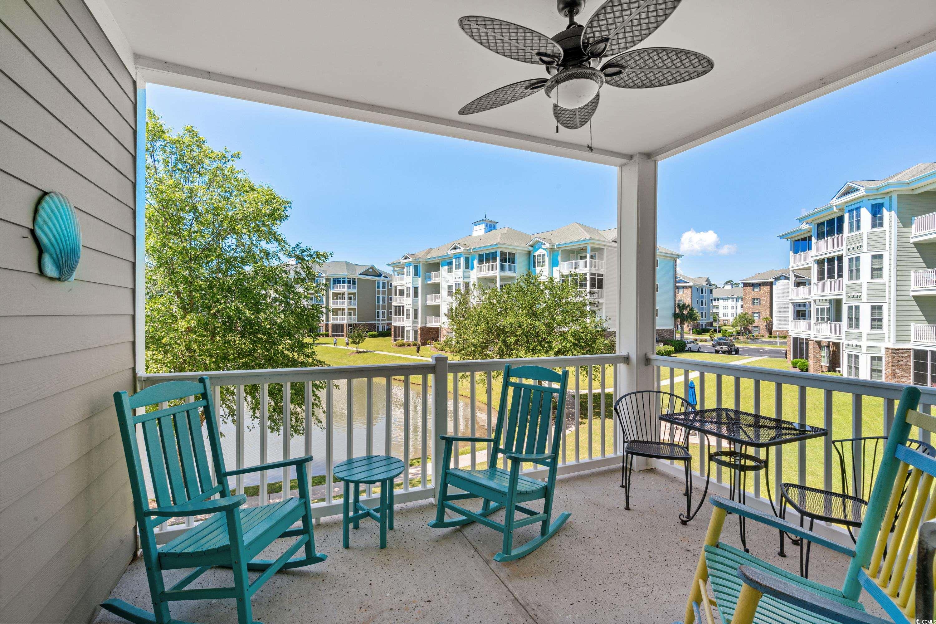 Covered balcony with turquoise rocking chairs overlooking a pond and neighboring apartment buildings.