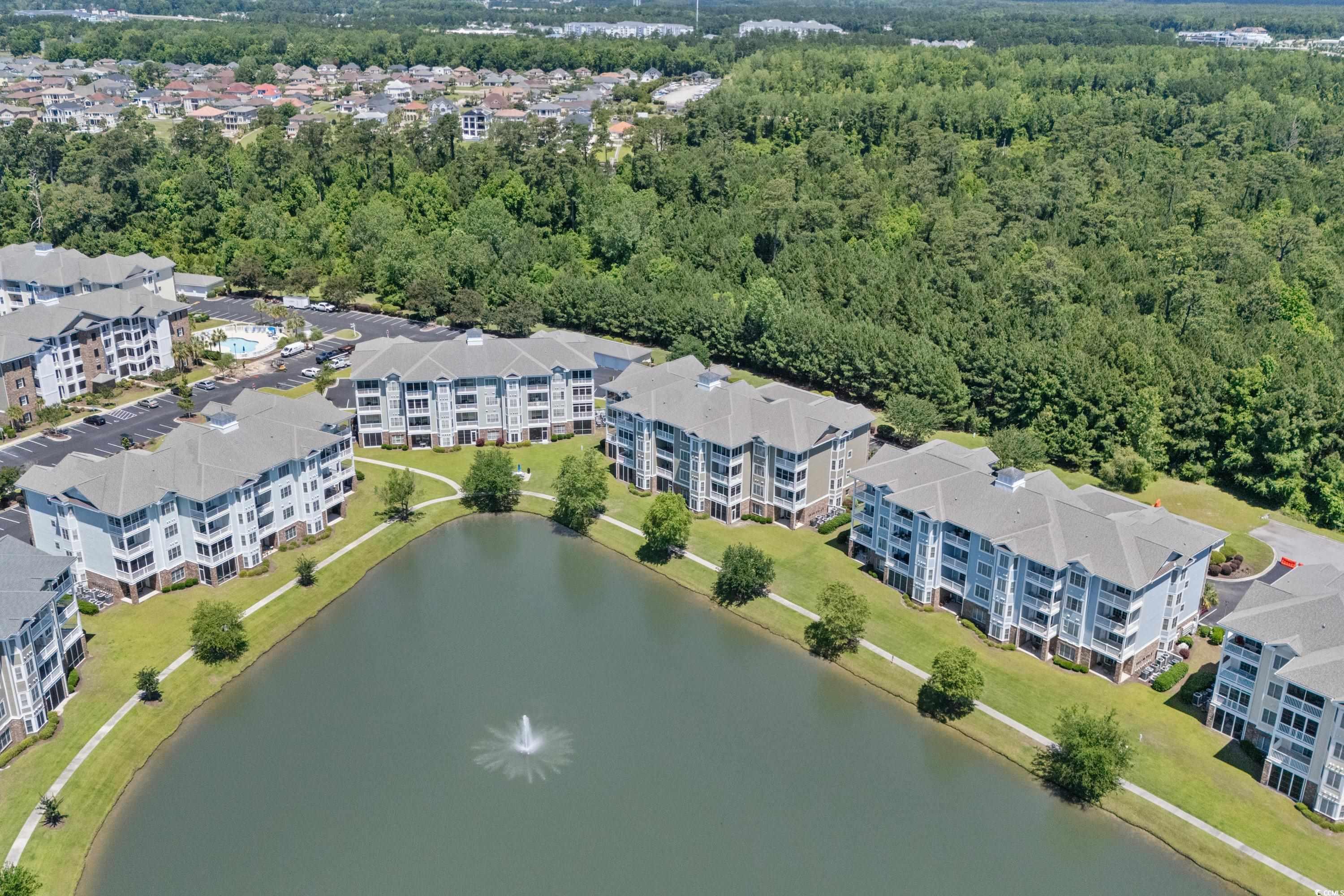 Aerial view of condominium buildings around a pond with a fountain and walking paths.