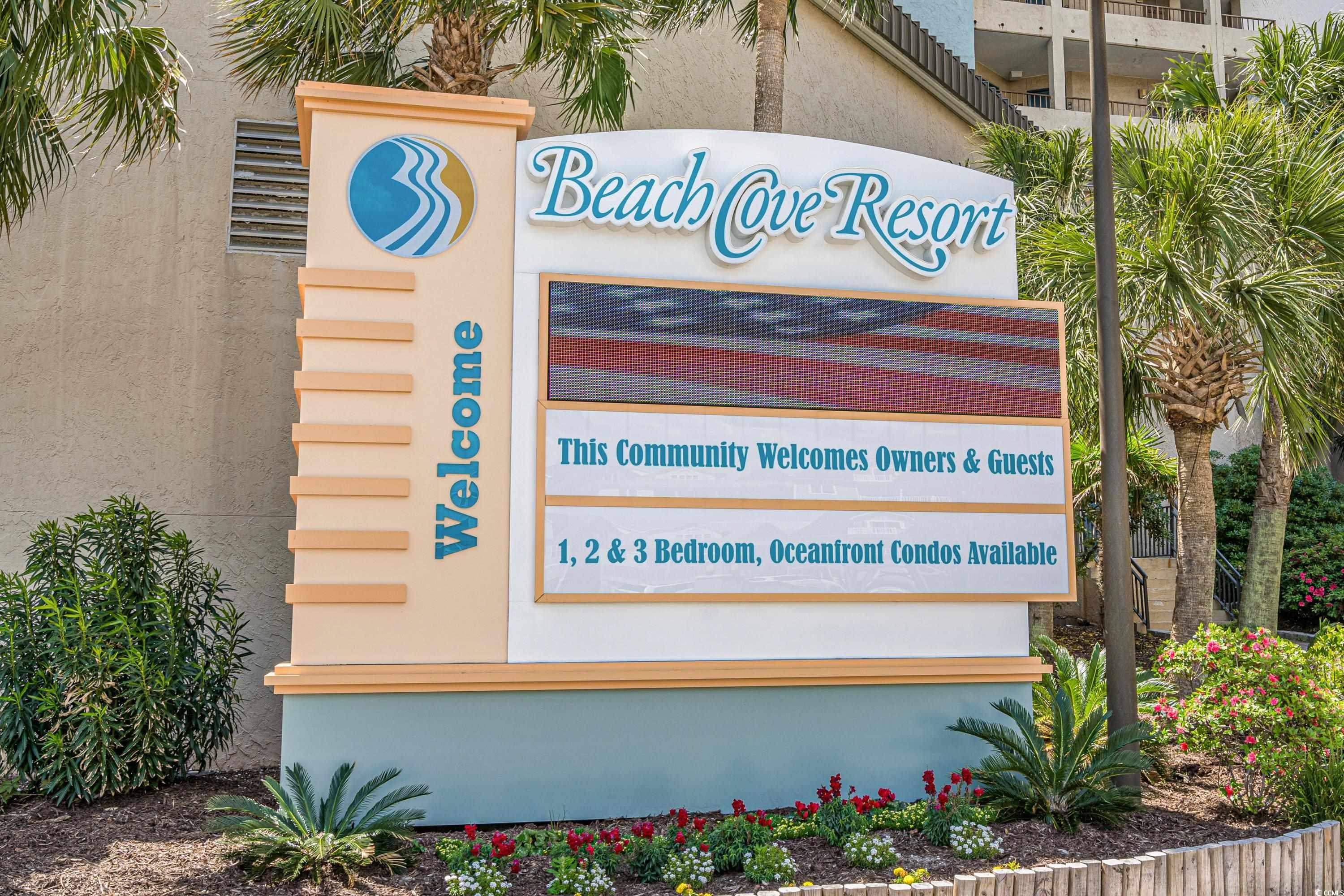 Beach Cove Resort welcome sign beside the condominium building, framed by palms and flower beds.