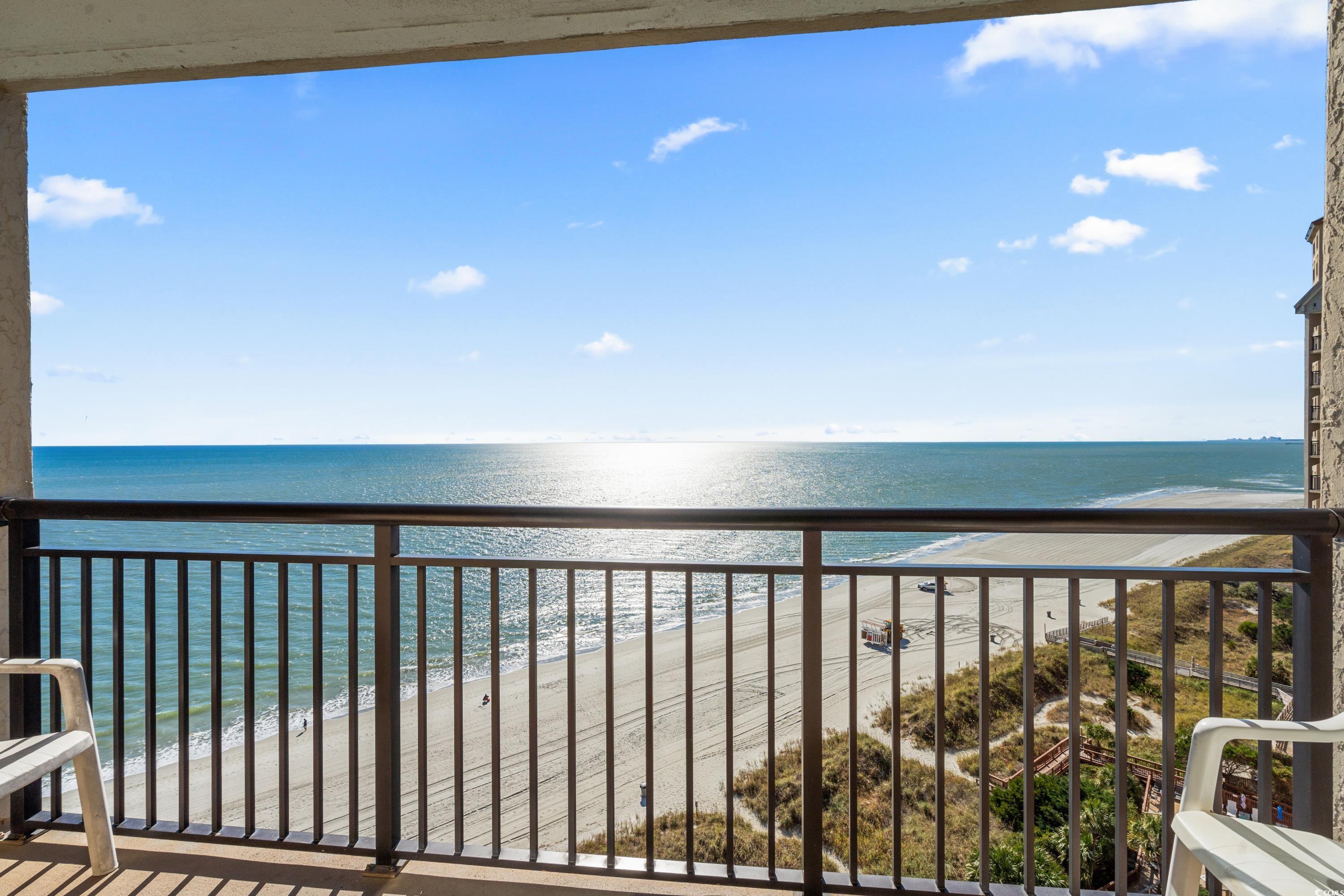 Oceanfront balcony overlooking the beach and blue water, framed by railings and patio chairs.