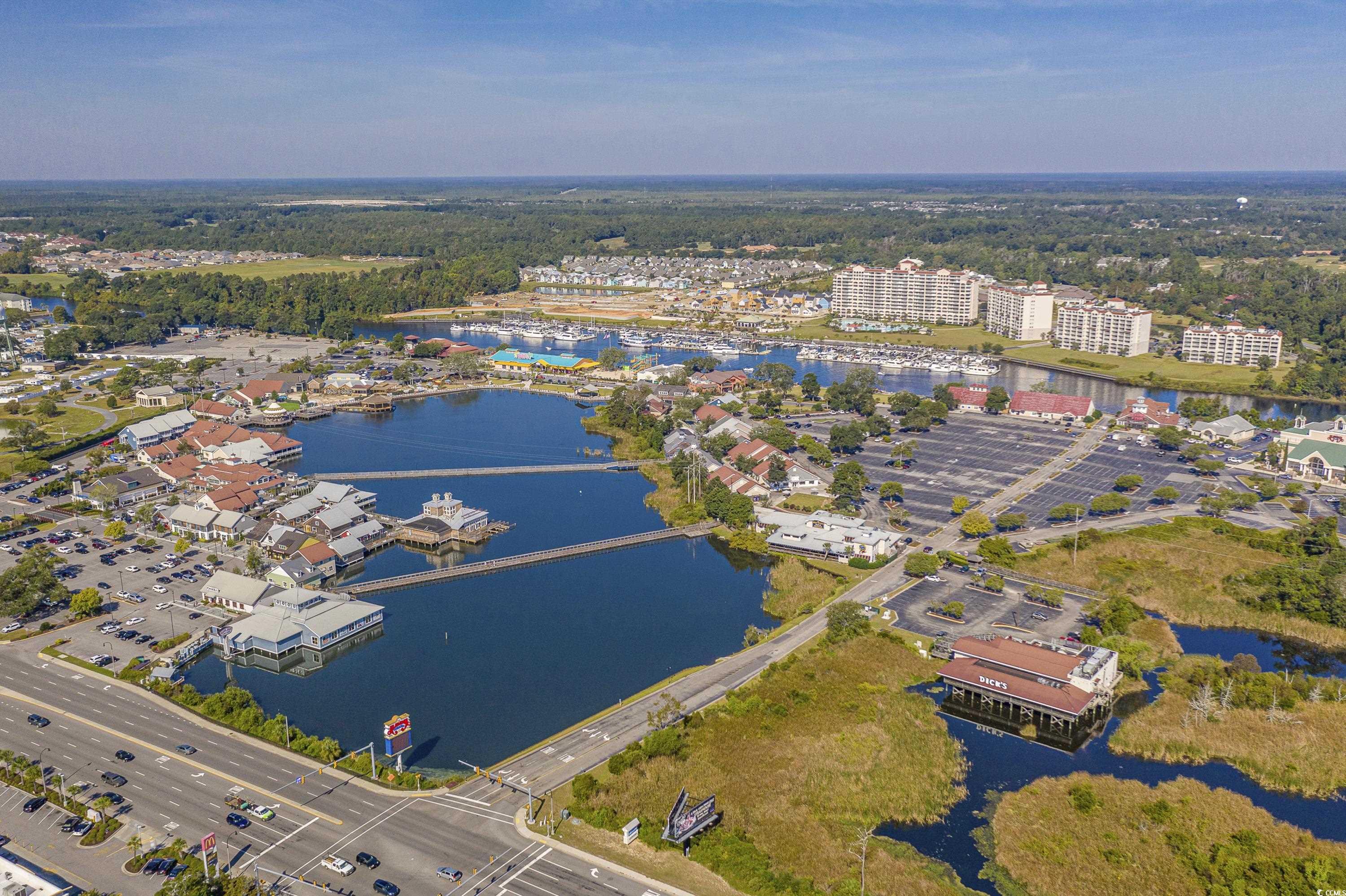 Aerial view of waterfront condos and marina beside shops, waterways, and a busy highway.