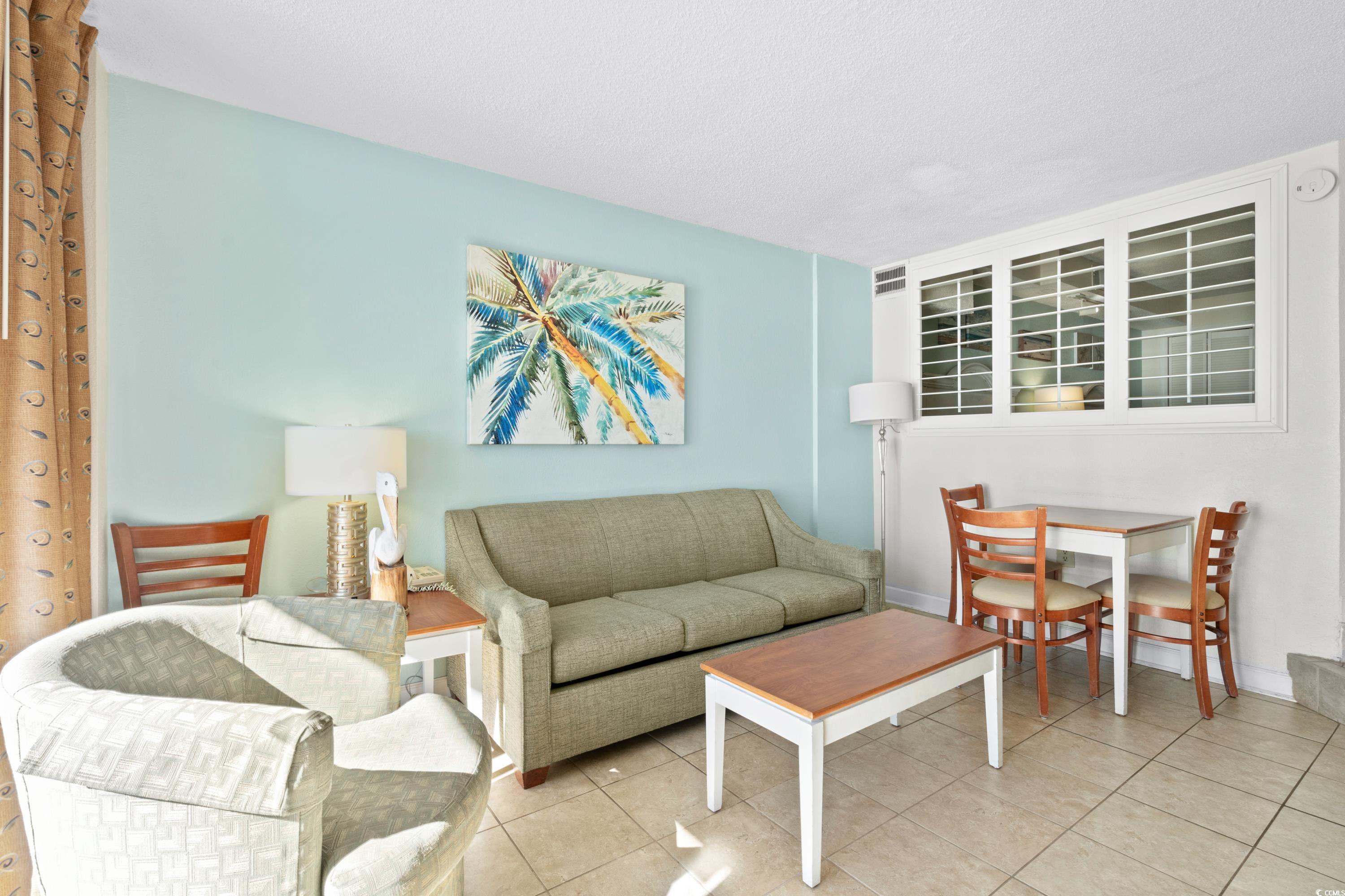 Condo living room with a sage-green sofa, pale armchairs, palm tree art, and an adjoining dining area.