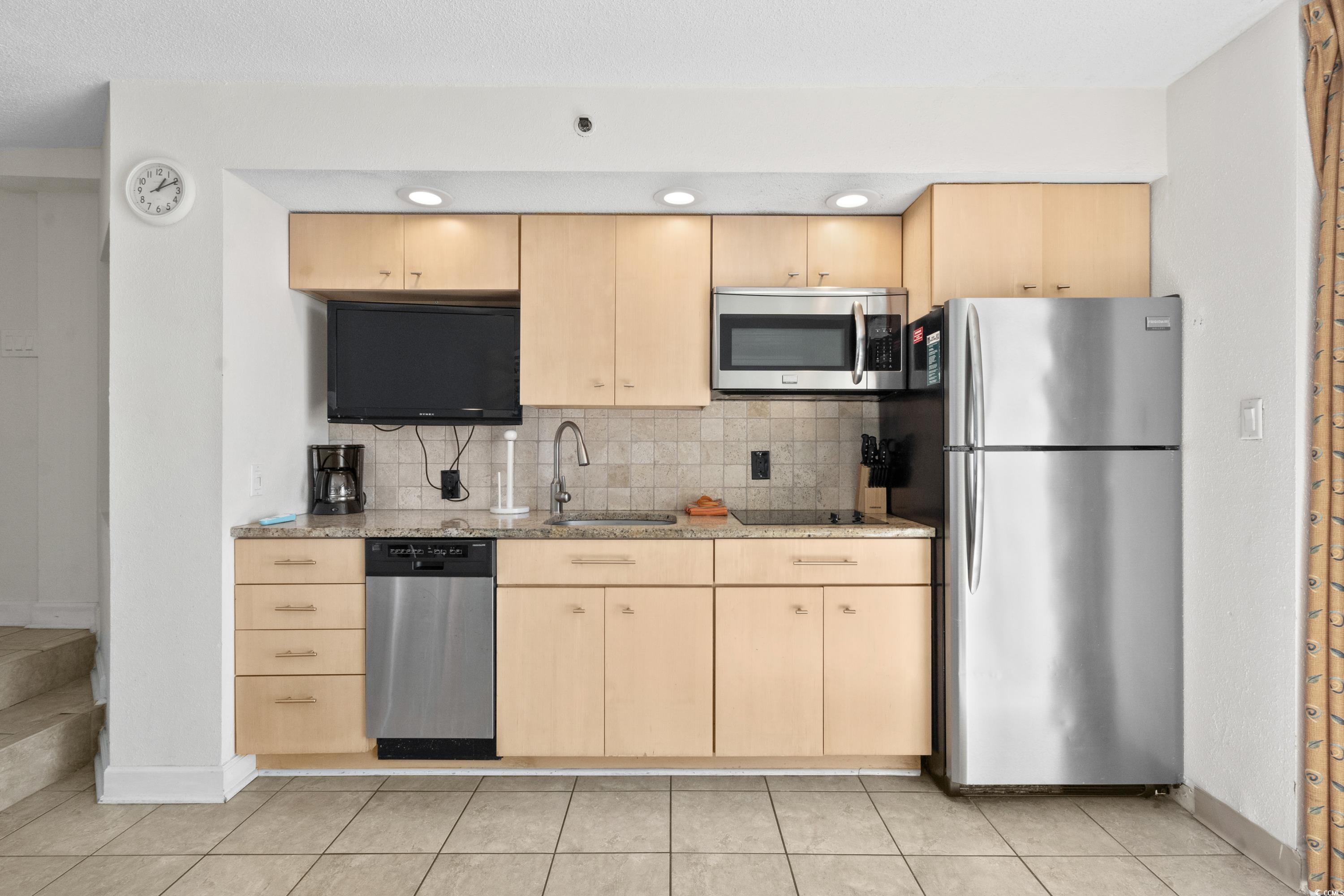 Condo kitchen with light wood cabinetry, stainless steel appliances, and a wall-mounted television.