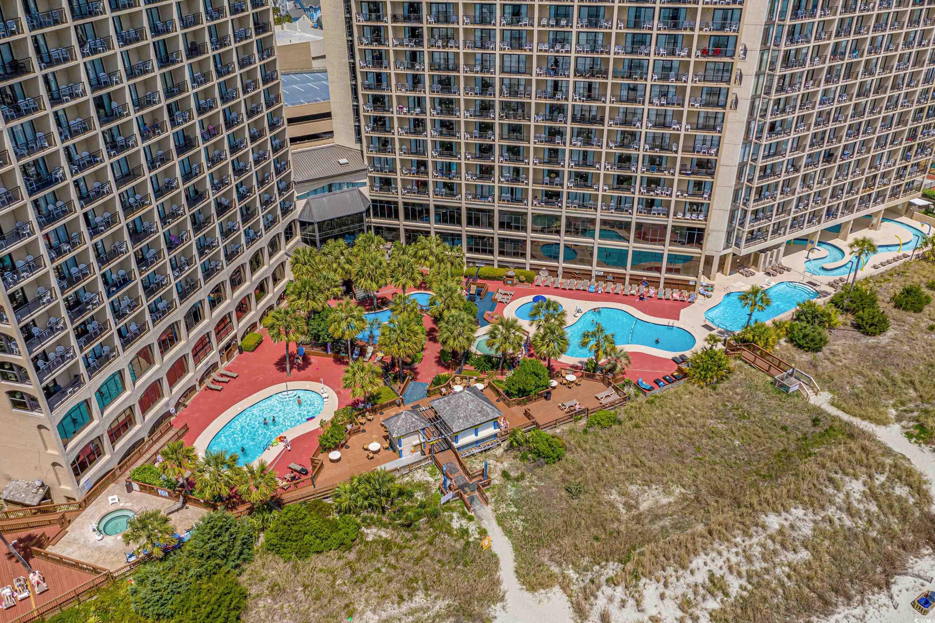 Aerial view of beachfront condo towers surrounding palm-lined pools and decks beside coastal dunes.