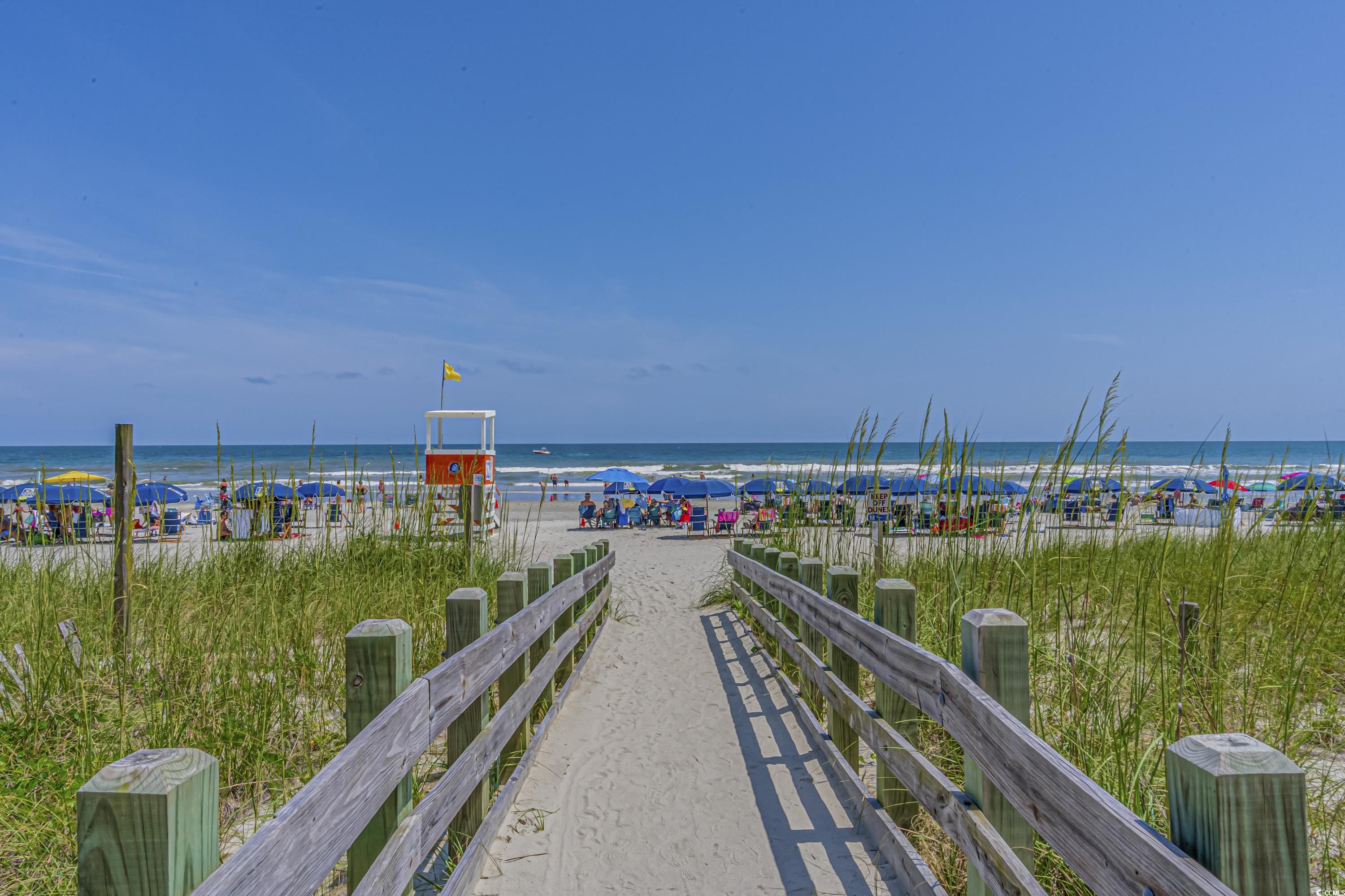 Beach access boardwalk crosses grassy dunes toward a crowded shore, with a lifeguard stand and ocean beyond.