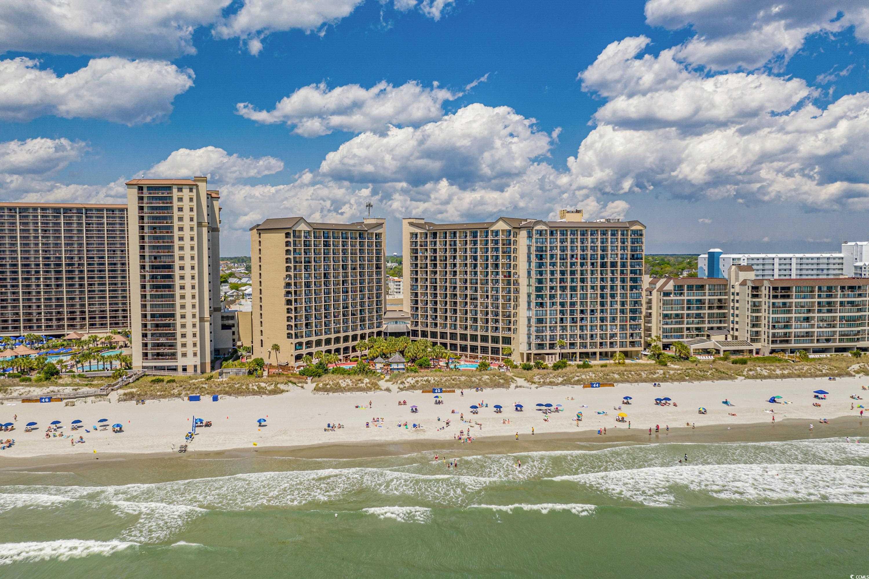 Oceanfront condominium towers above a busy sandy beach, seen from offshore with pools and waves.