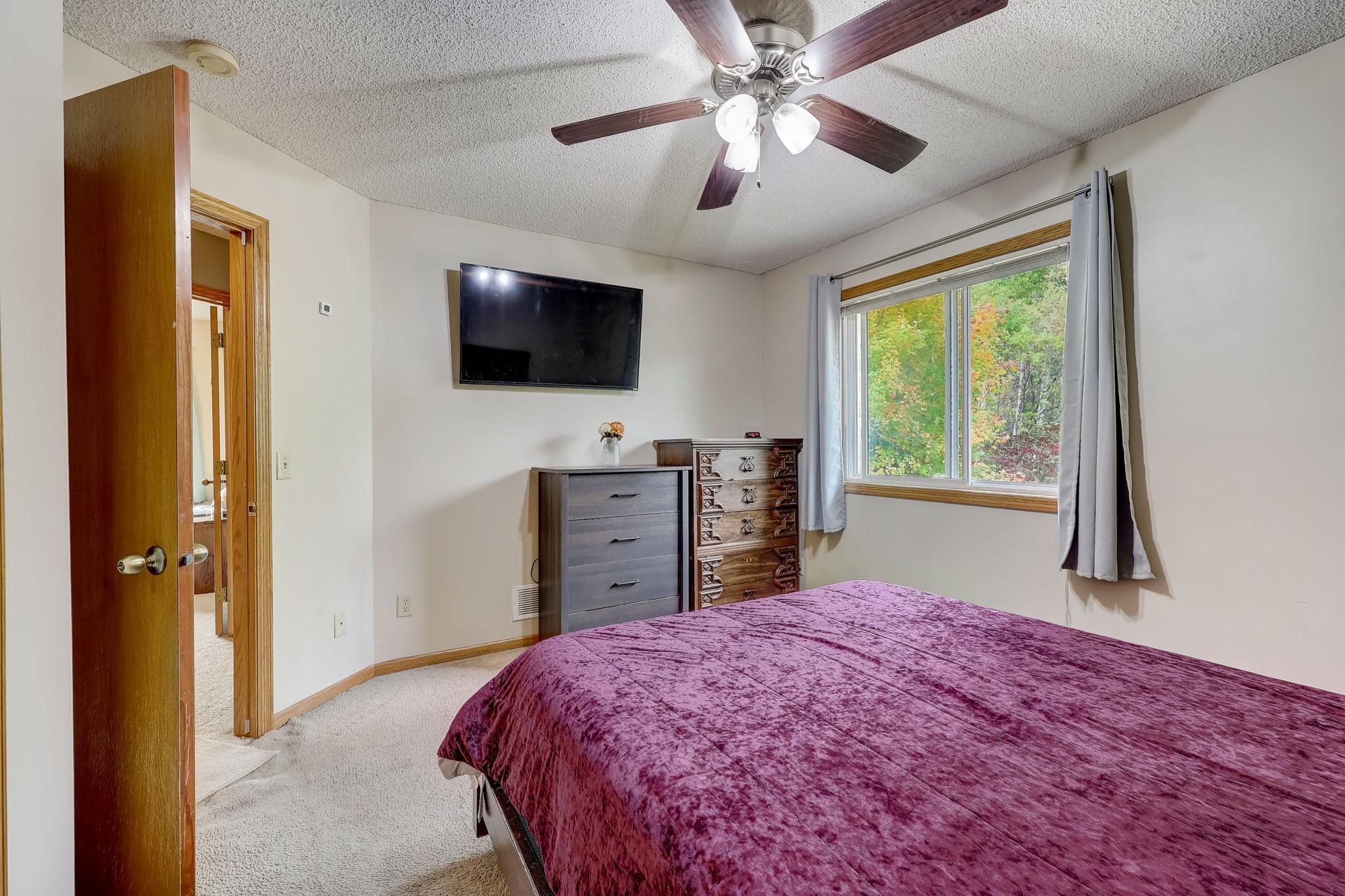 Bedroom with a burgundy bedspread, two dressers, and a window overlooking autumn trees.