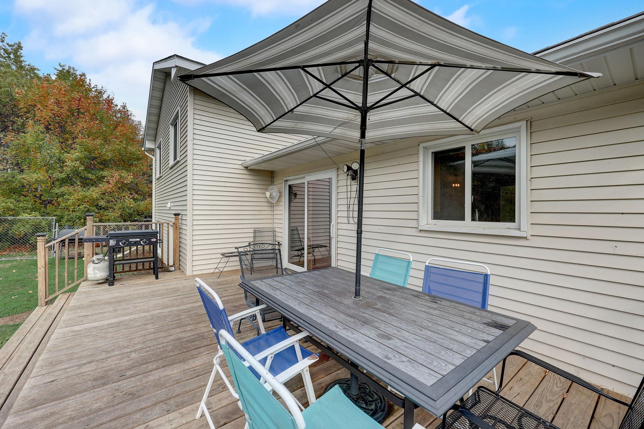 Wooden deck with patio dining set and large umbrella beside the house, with a grill and fenced yard.
