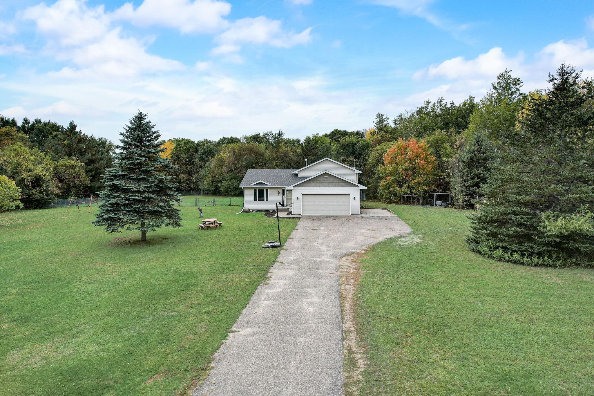 House with attached garage at the end of a long driveway, surrounded by a broad lawn and mature trees.