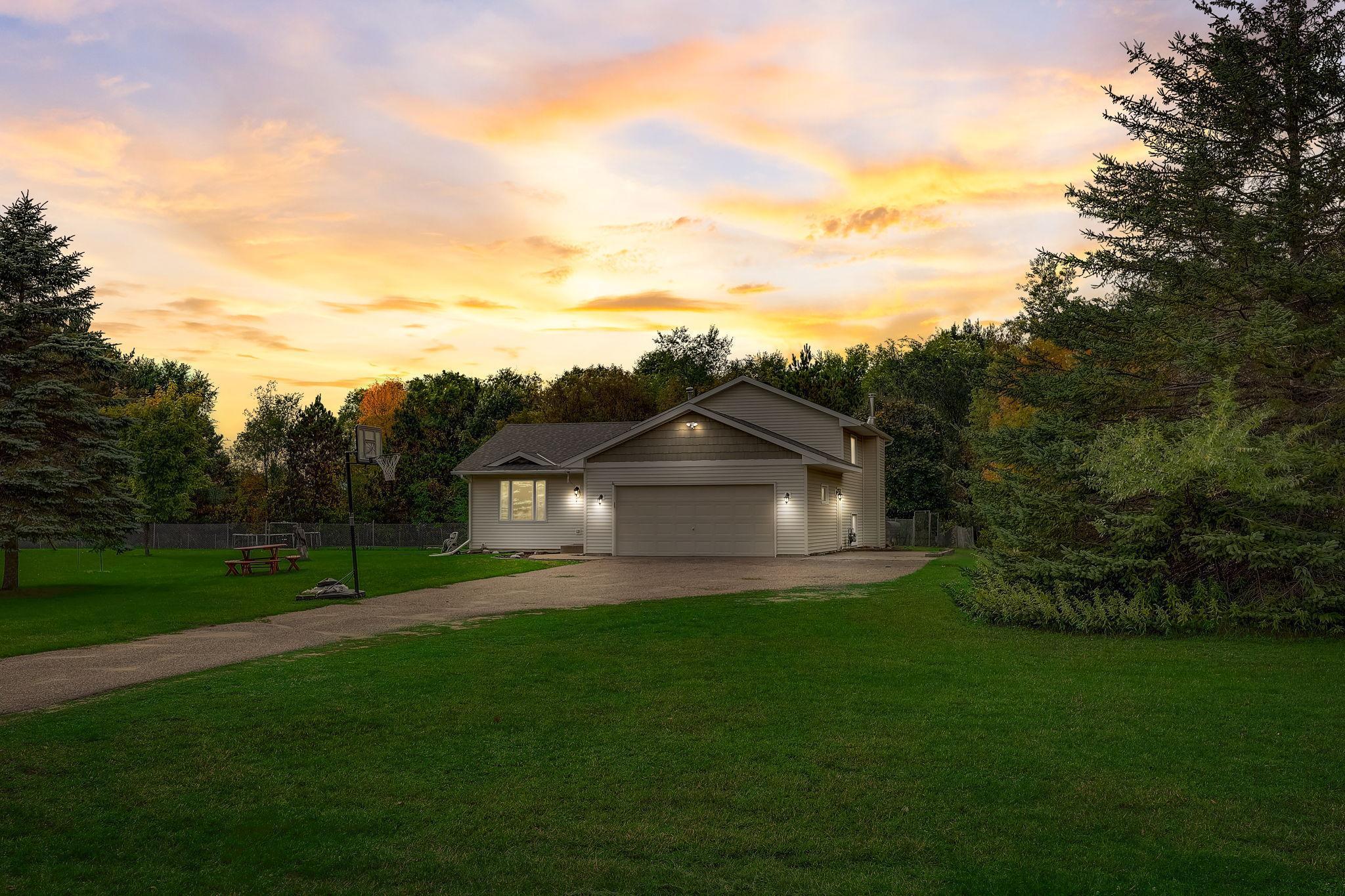 Two-story home with a double garage, broad lawn, basketball hoop, and trees at sunset.