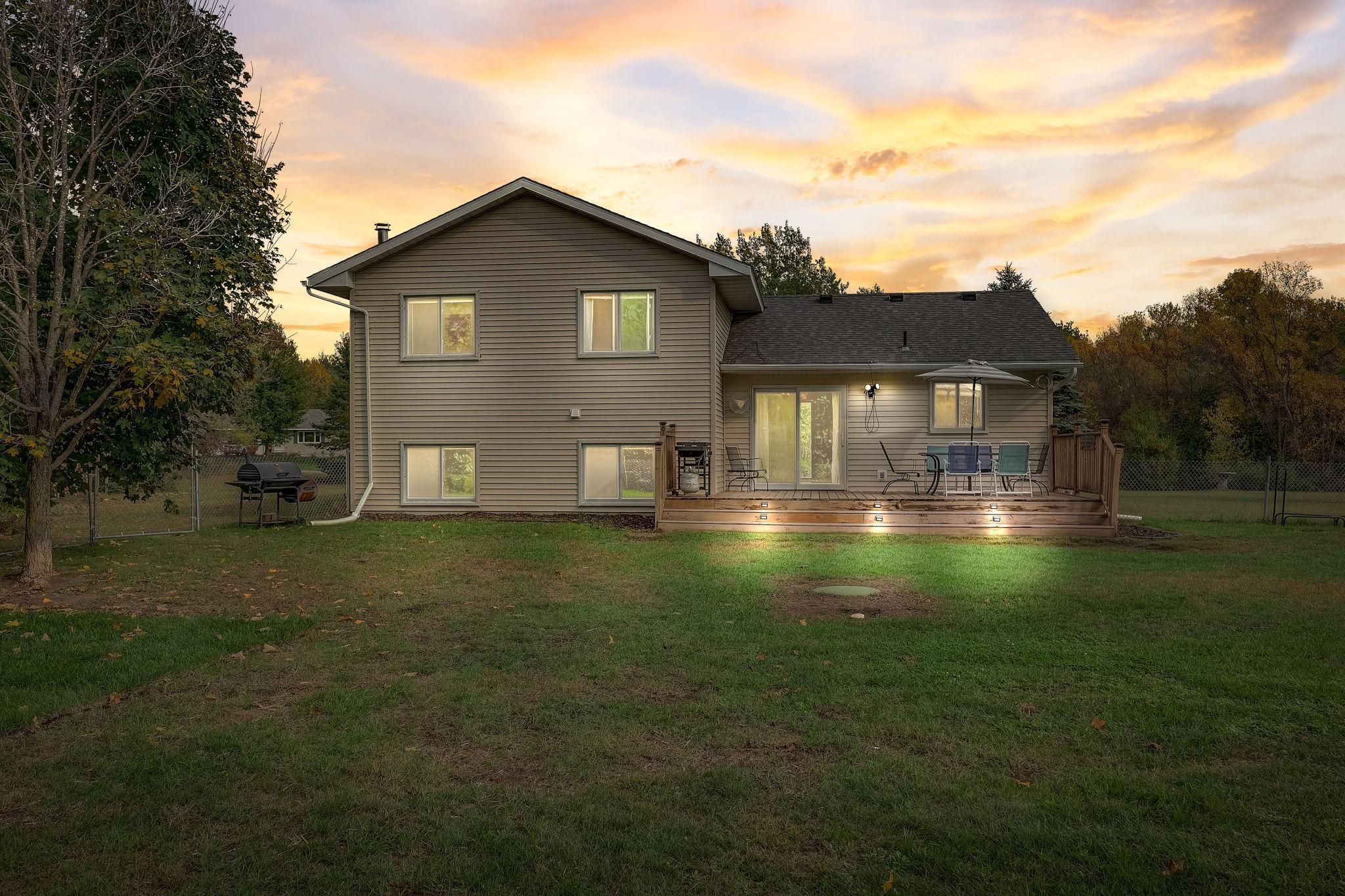 Home’s rear exterior at sunset, with a raised deck, patio furniture, and fenced lawn.